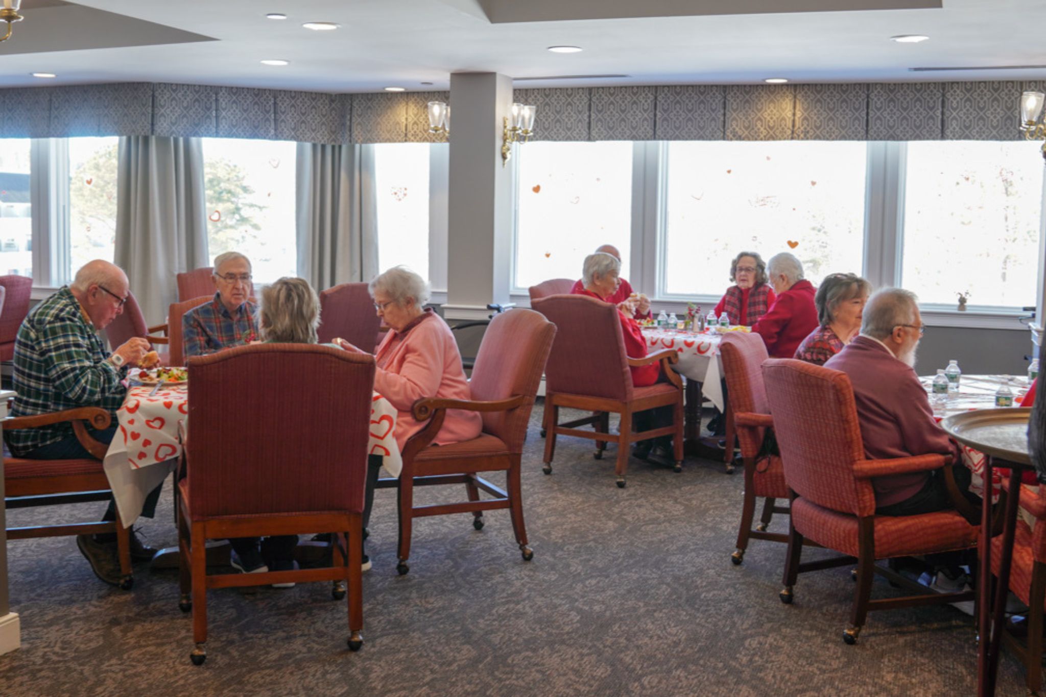 Elderly people, some sharing stories about their scholarship days, sit and eat together at round tables in a bright, decorated dining room.
