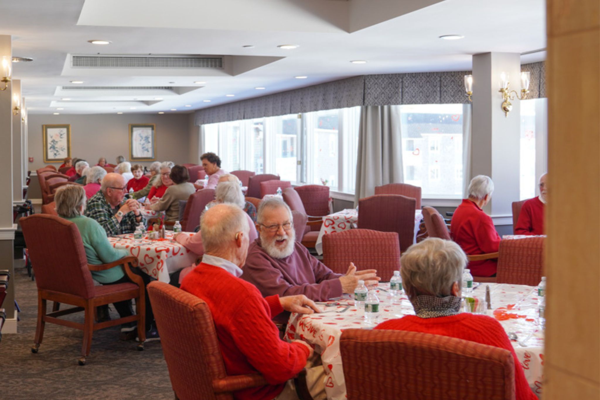 Elderly people sit at decorated tables, sharing stories about scholarship and life experiences as they dine together in a bright, communal dining room.