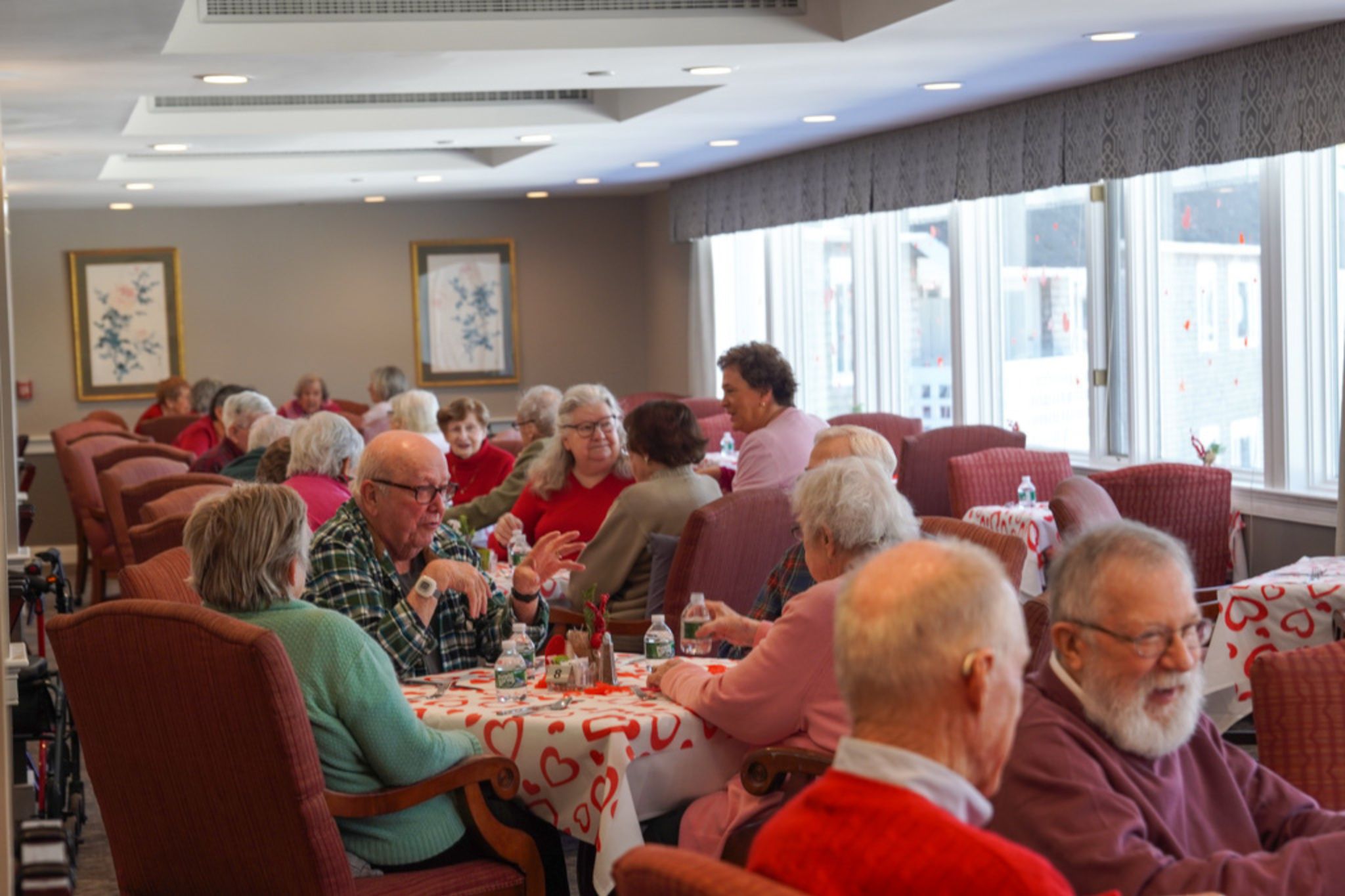 Elderly people sit at tables with heart-themed tablecloths, chatting about friendship and scholarship opportunities in a bright, lively room.