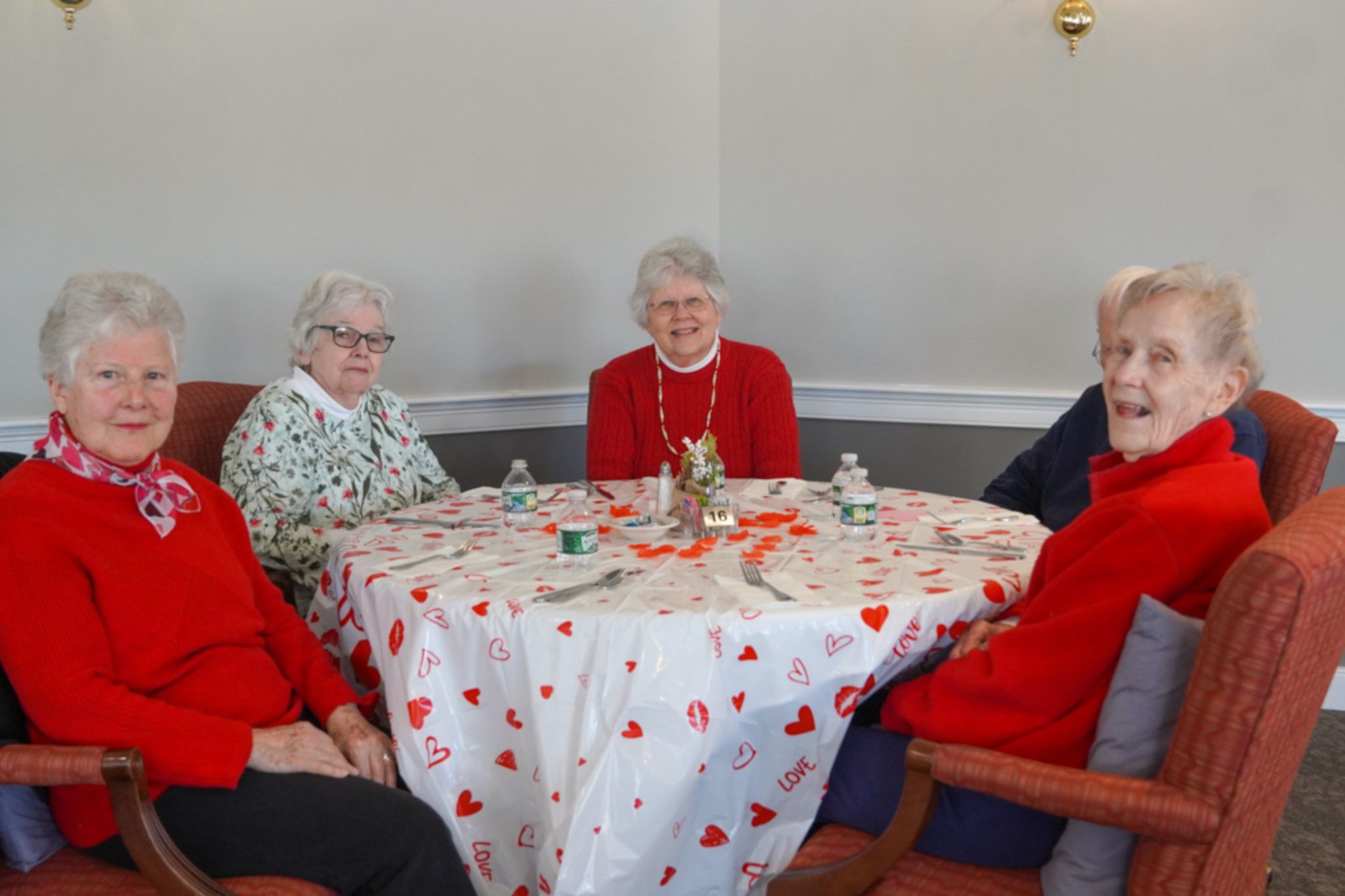 Five elderly women sit around a heart-themed table, smiling and enjoying each other's company as they share stories about friendship and a recent scholarship awarded in their community.