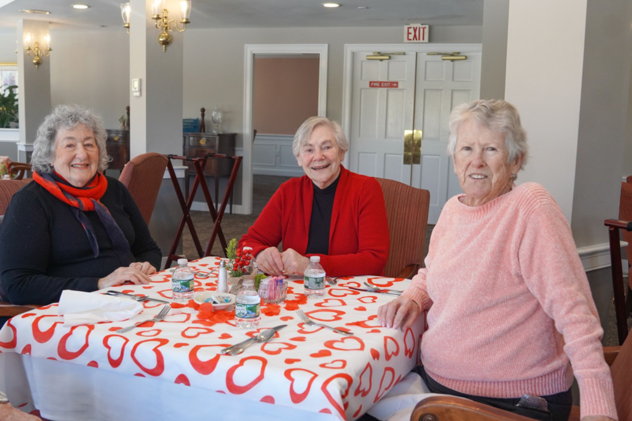 Three older women sit smiling at a table with a heart-patterned tablecloth, bottled water, and a brochure about a local scholarship.