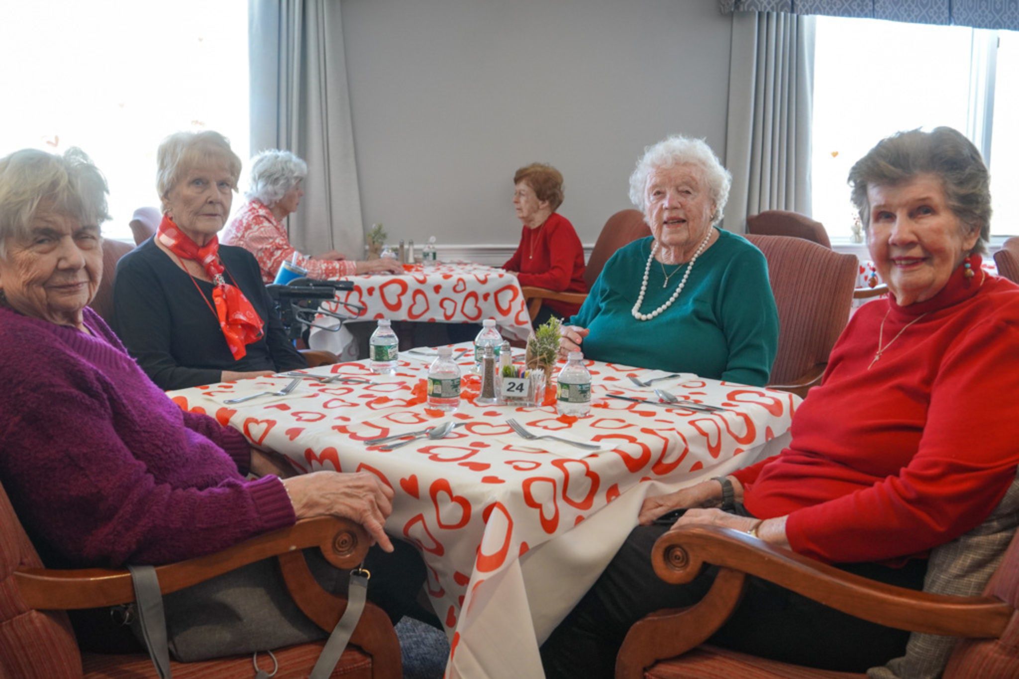 Four elderly women sit at a table with a heart-patterned tablecloth in a bright, cozy dining room, sharing laughter and stories of friendship and scholarship over tea.