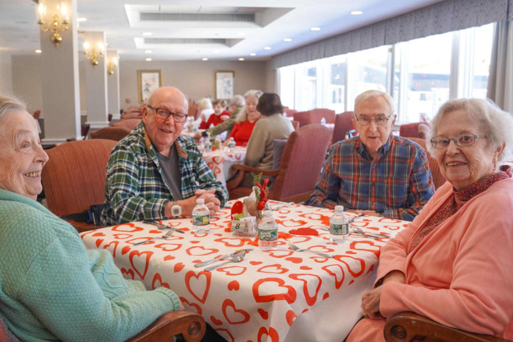 Four smiling seniors sit at a heart-decorated table in a bright, cheerful dining room, sharing stories about their lives and scholarship achievements.