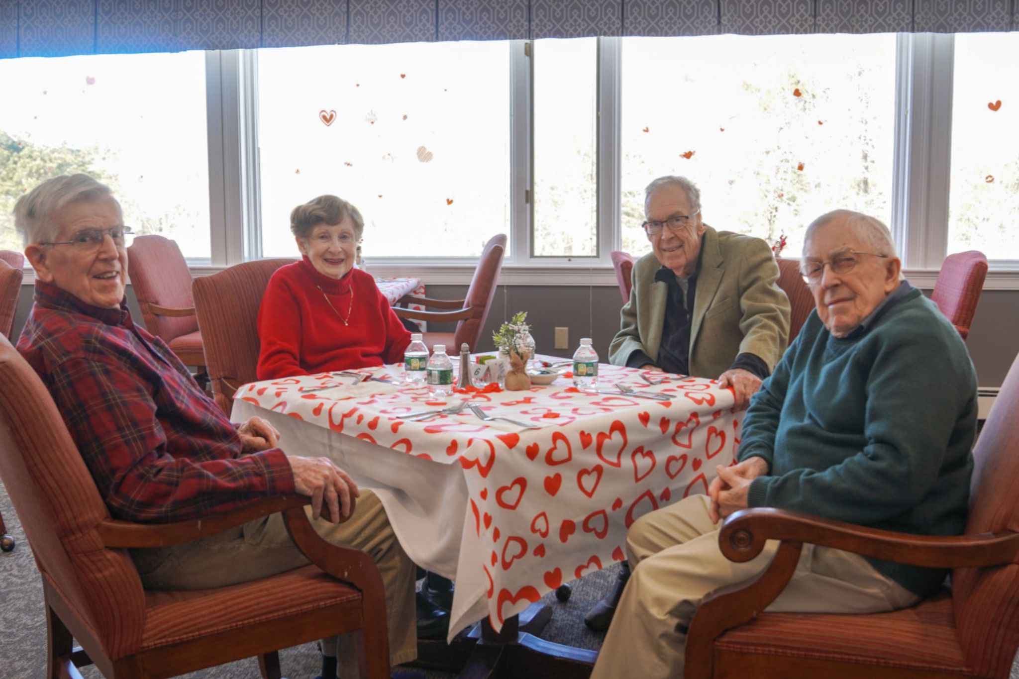 Four smiling seniors sit around a table with a heart-patterned tablecloth in a bright, cozy room, sharing stories about their experiences with scholarship opportunities.