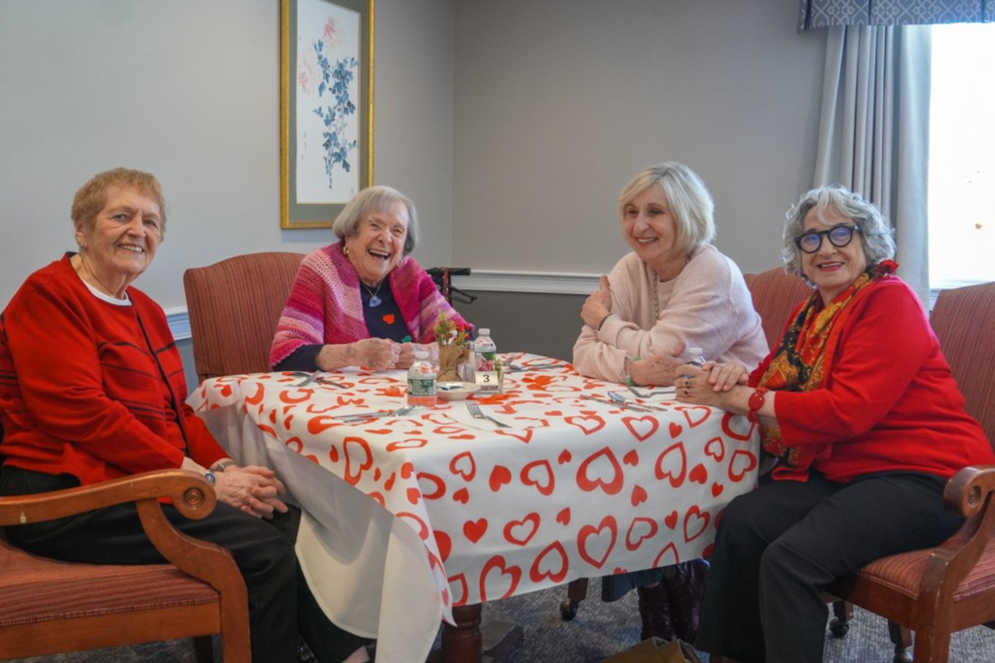 Four older women, smiling and sitting around a heart-patterned table in a cozy room, discuss scholarship opportunities over tea.