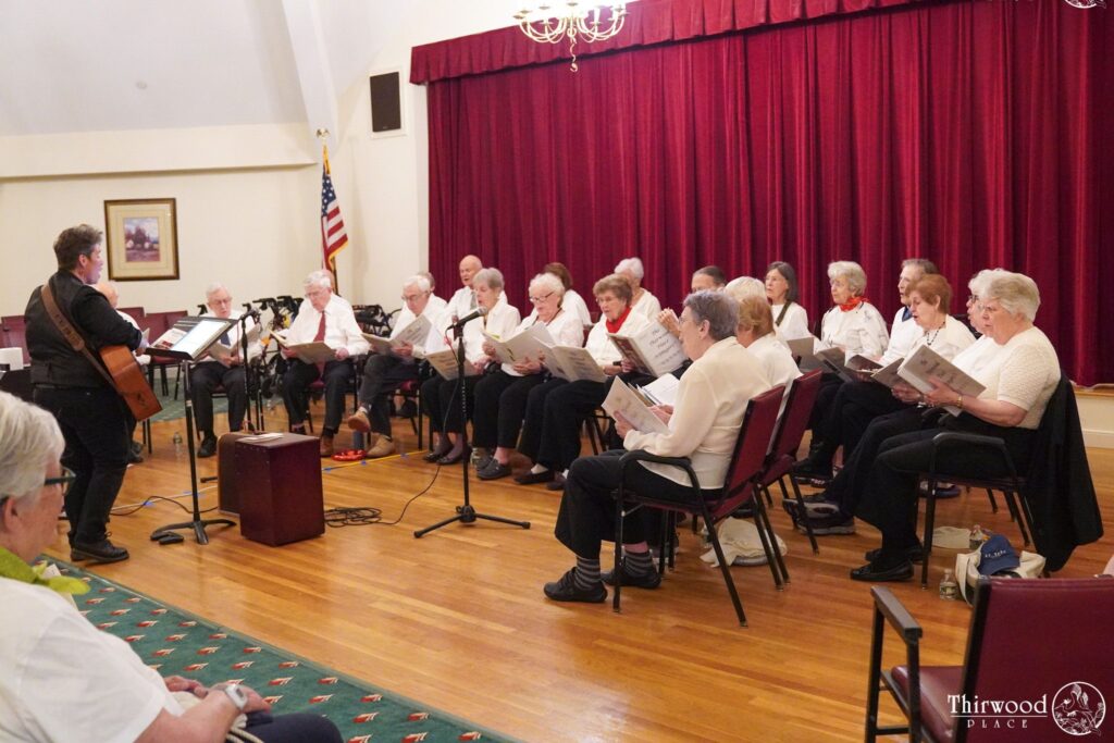 A group of seniors sing in a choir with a guitarist in a room with red curtains and microphones, celebrating their shared passion for music through a scholarship-supported program.