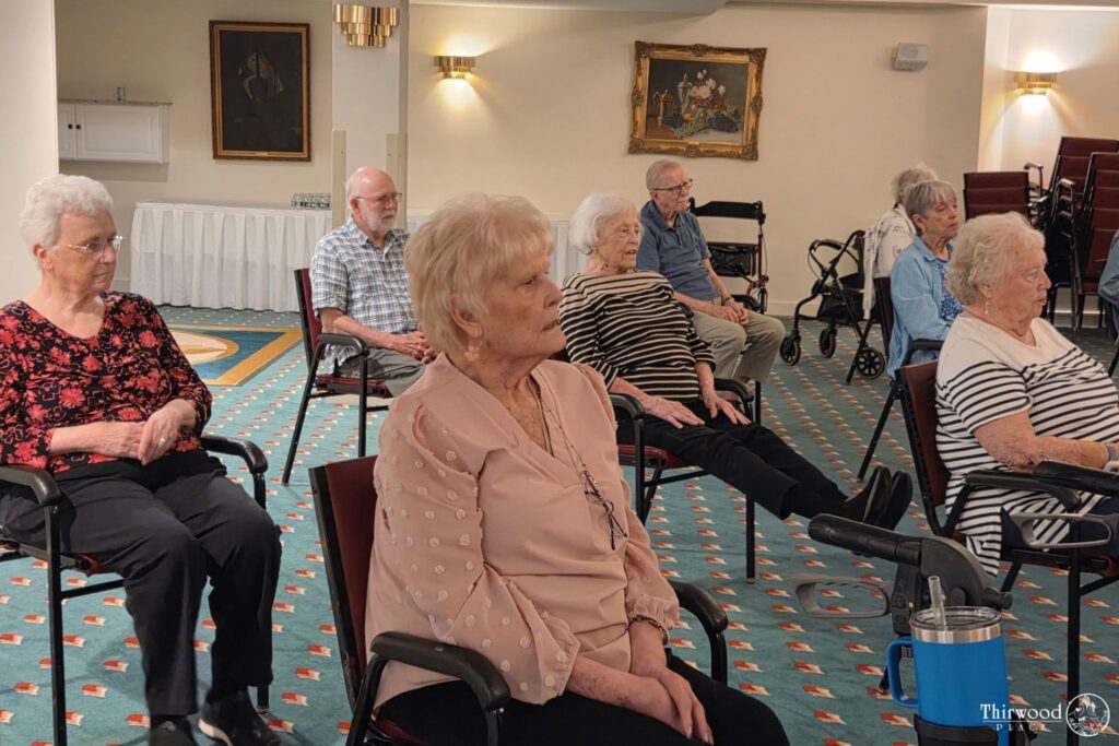 A group of seniors in chairs participates in a seated exercise class in a carpeted room, with some discussing a local scholarship program during breaks.