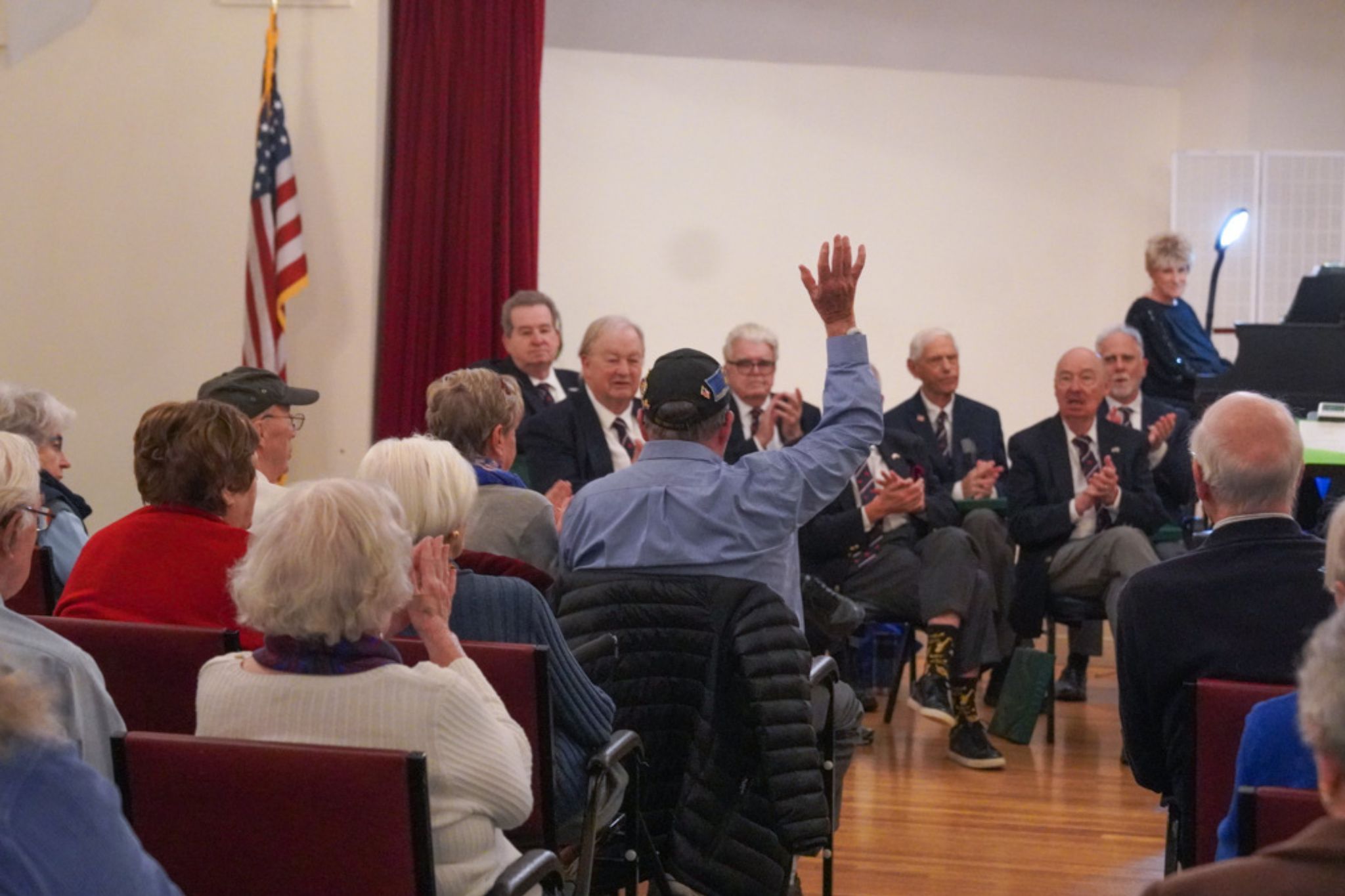 Older adults seated in a hall; one person raises a hand while others applaud, celebrating a scholarship. U.S. flag in the background.