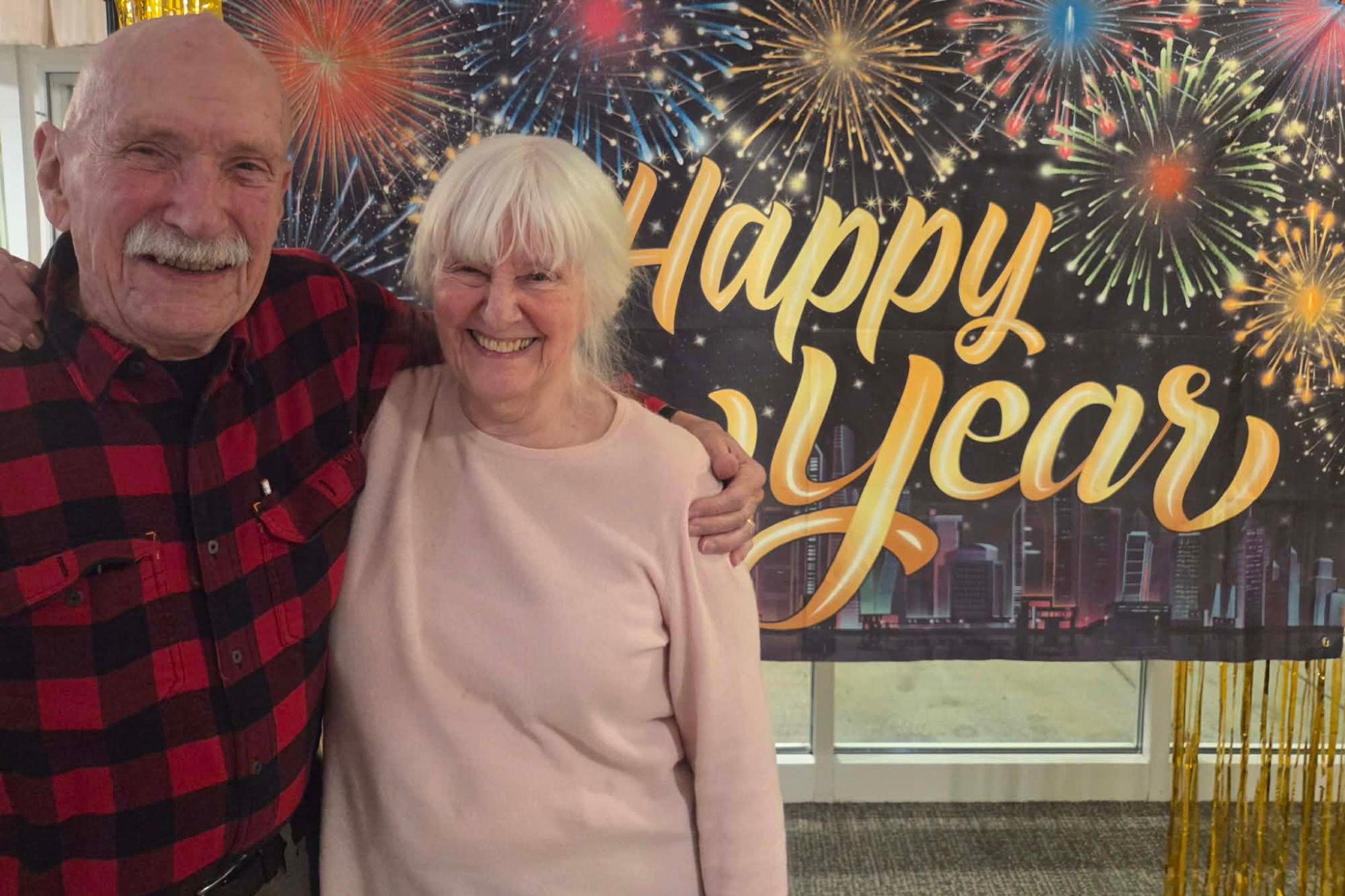 Smiling elderly couple stands in front of a Happy New Year banner with fireworks in the background, capturing a joyful moment perfect for a photo essay about summer celebrations and timeless happiness.