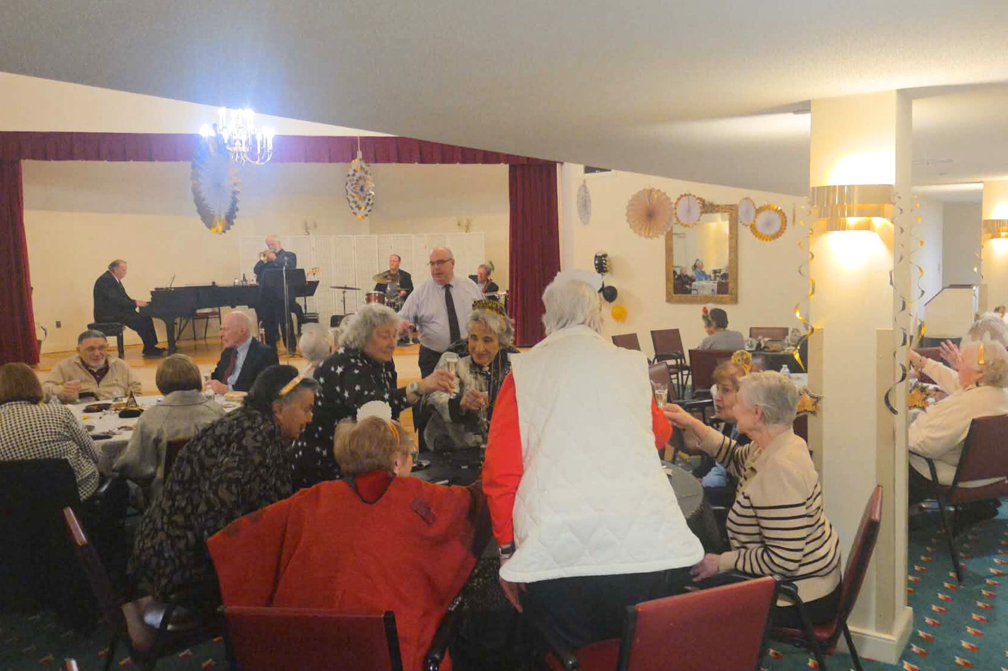 A group of seniors socialize at round tables while a pianist performs onstage at a decorated event hall, capturing the warmth and joy of a summer gathering perfect for a photo essay.