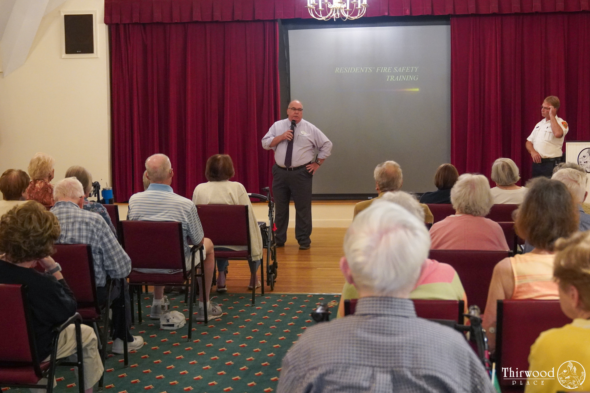 A man speaks to seated seniors during a fire safety training session in a community room, also sharing tips on how to avoid cold and flu and what to do if someone begins to choke.