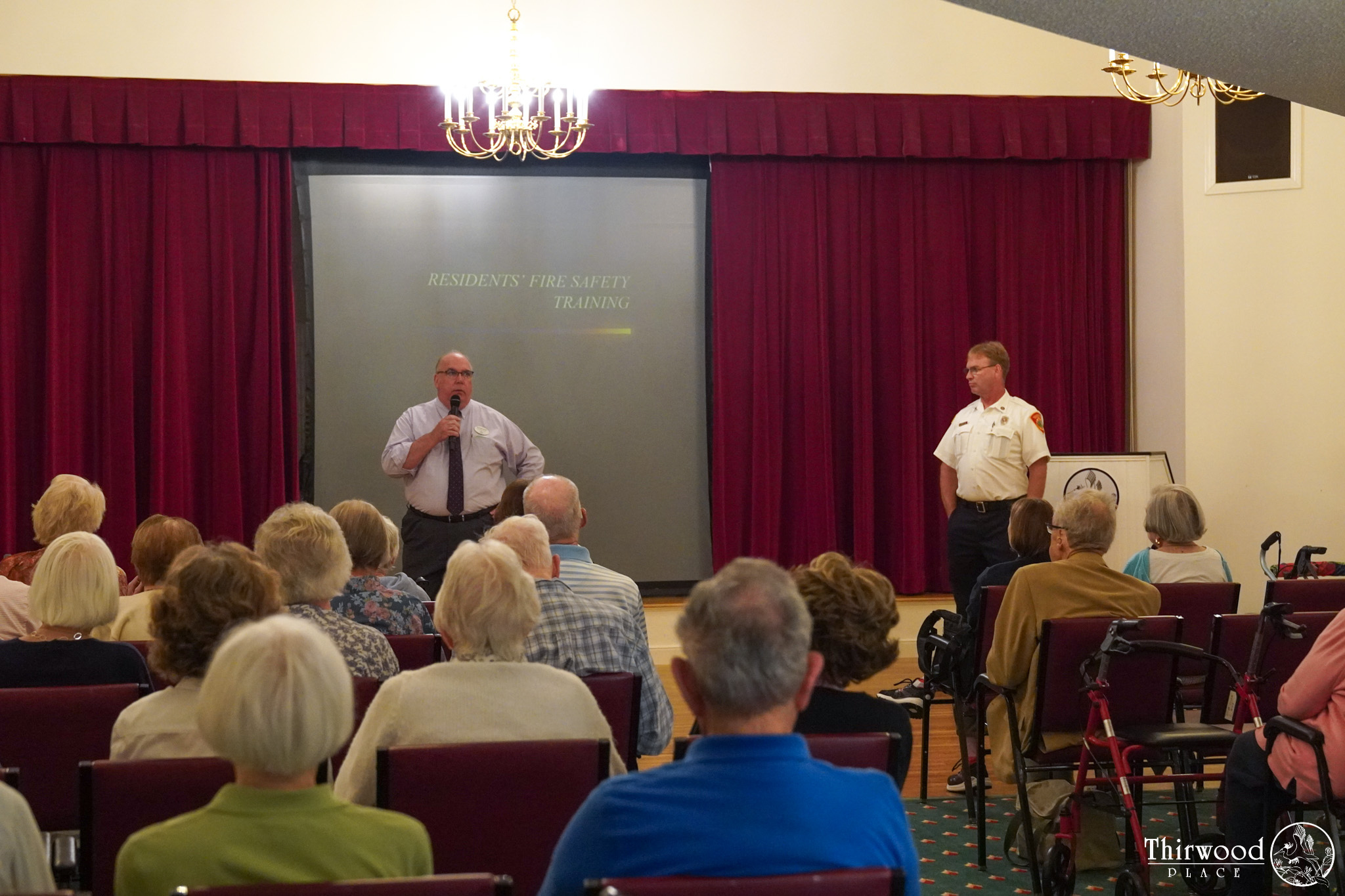 Two men speak to a seated audience about fire safety training, discussing what to do if someone begins to choke, in a room with red curtains and chandeliers.