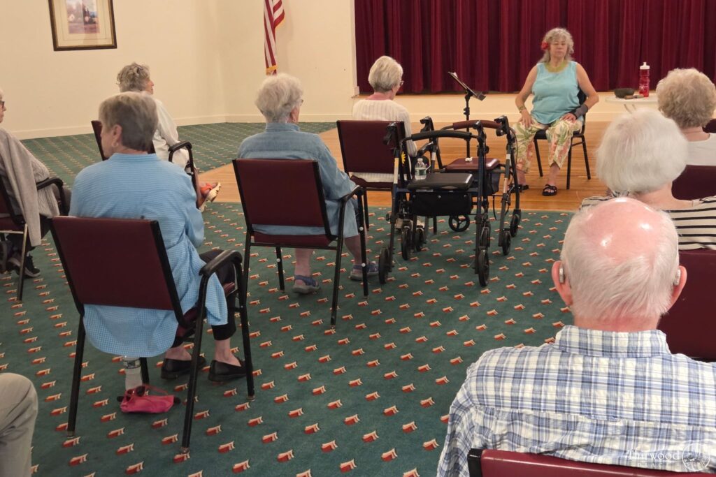 A group of seniors sit facing a woman speaking on stage about a scholarship program in a community room with red curtains.