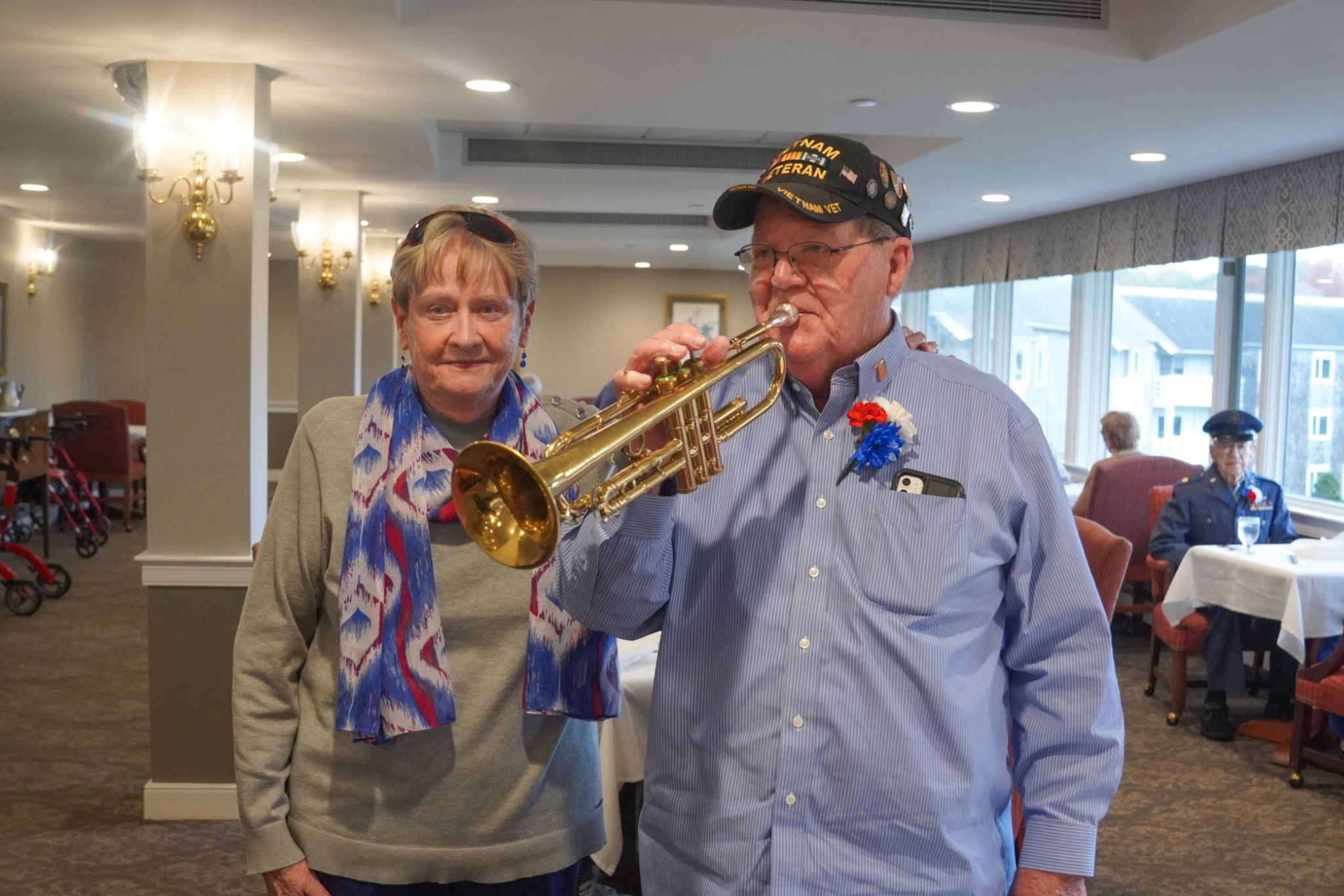 Older man in a veteran cap plays trumpet while a woman beside him smiles, celebrating his scholarship award in a well-lit room.