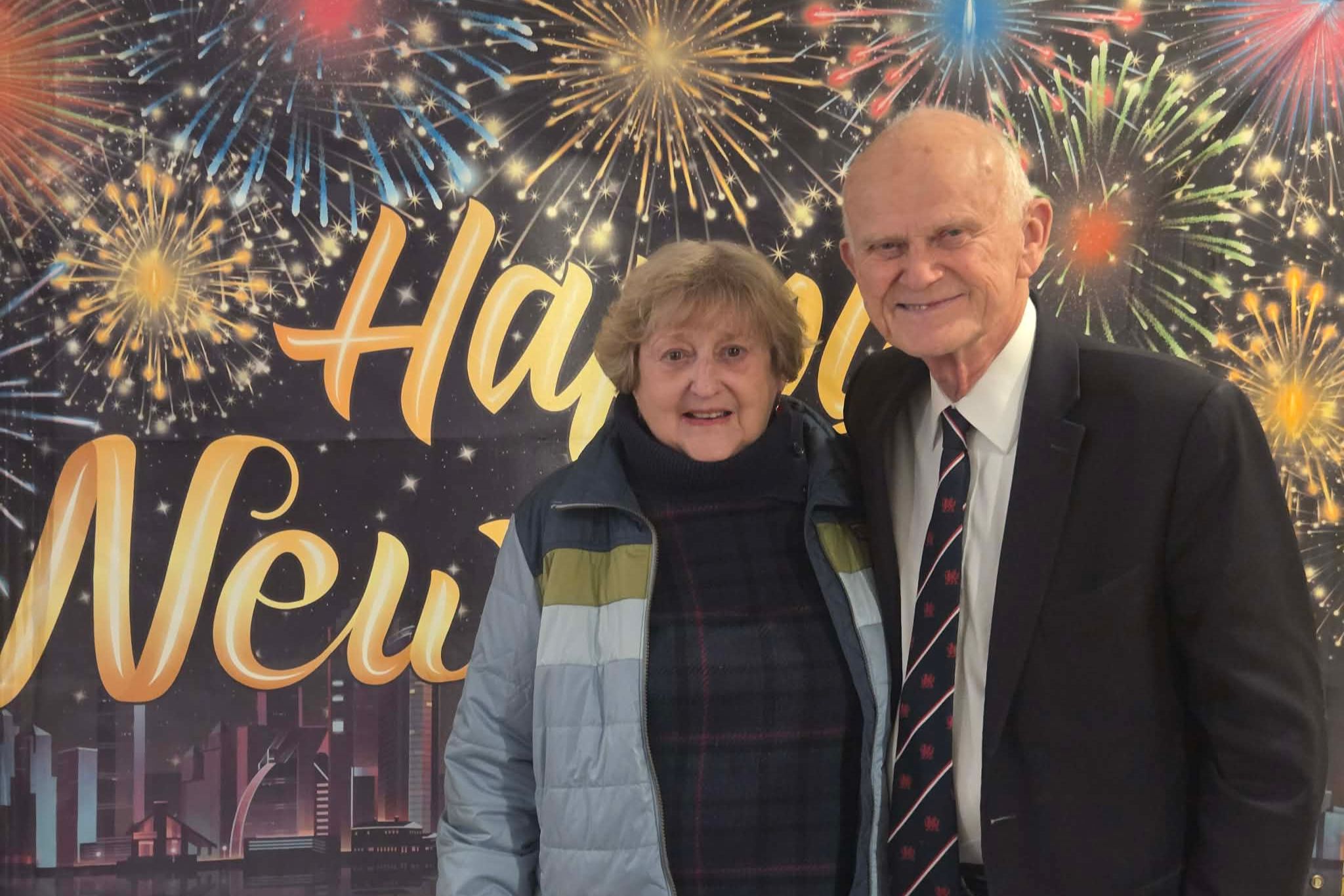 Older couple smiling in front of a Happy New Year sign with festive fireworks in the background, perfect for a photo essay capturing joyful summer celebrations.