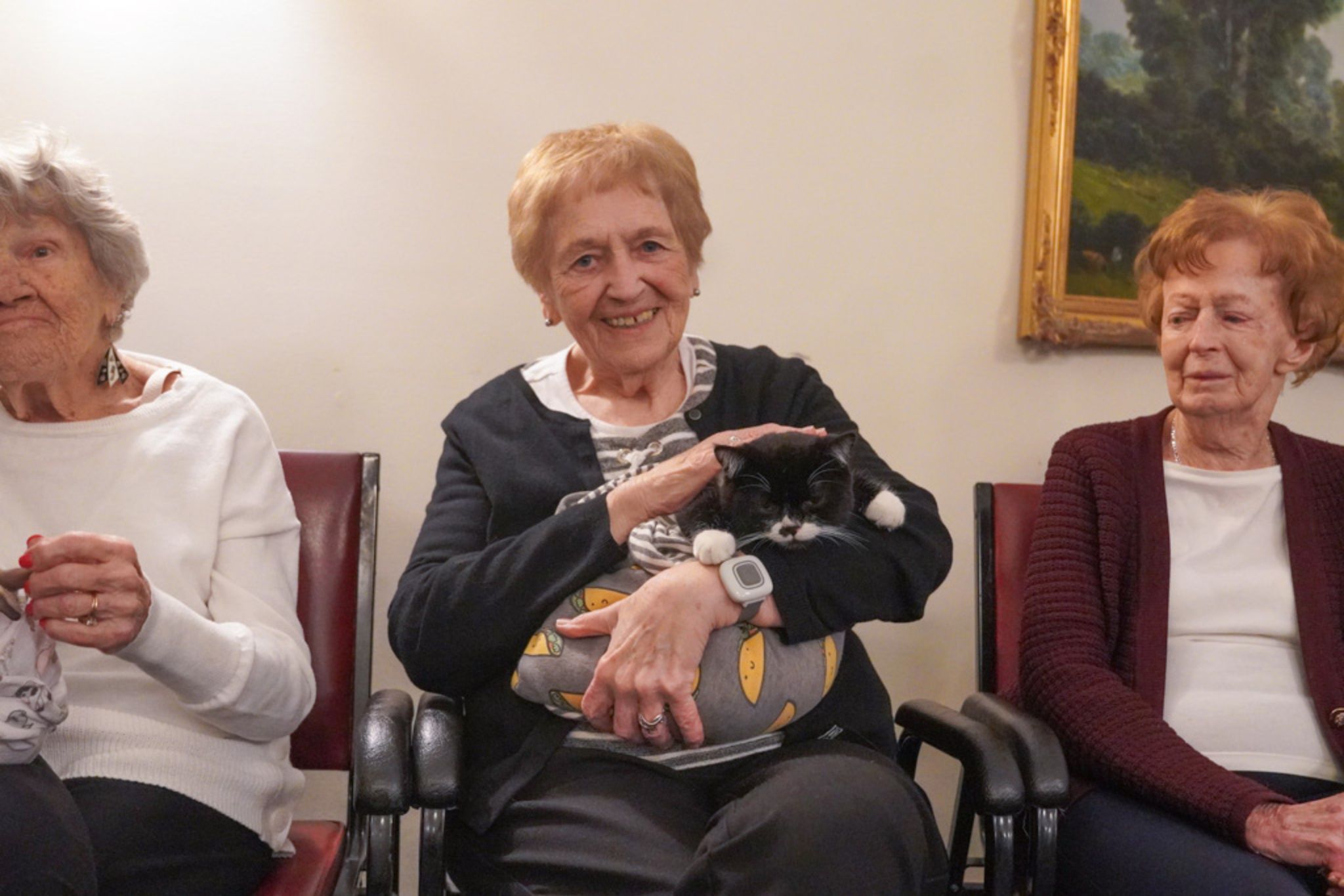 Three elderly women sit together; the middle woman is smiling and holding a black and white cat on her lap, perhaps sharing stories of friendship or celebrating a scholarship earned by someone dear.