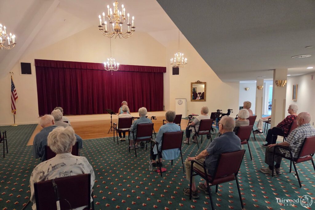 Elderly people seated in a room facing a stage with red curtains, attentively watching a speaker discuss scholarship opportunities or perform.