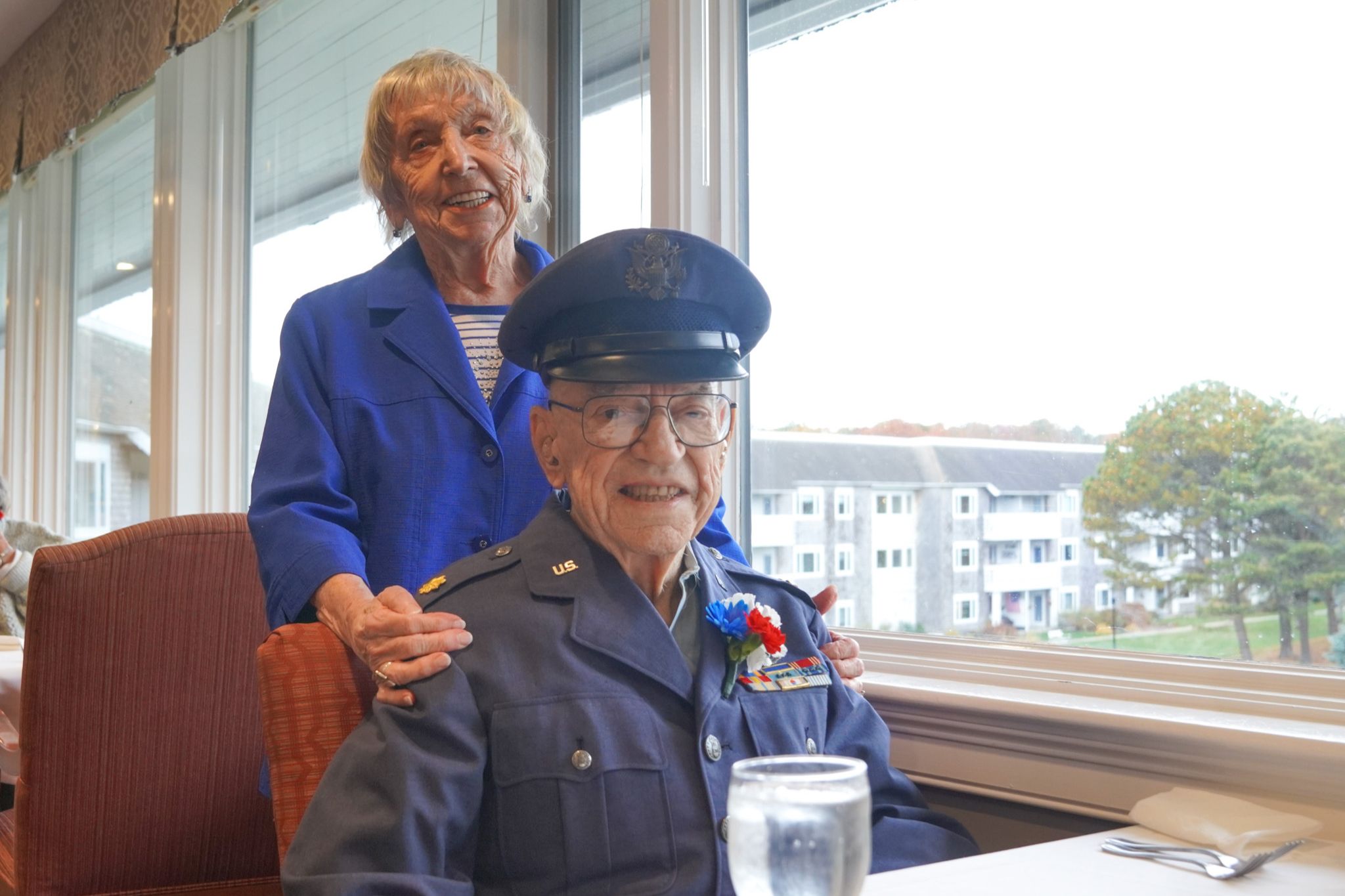 Smiling elderly man in military uniform sits at a table, proudly displaying his scholarship certificate, with a smiling woman standing behind him inside a bright room.