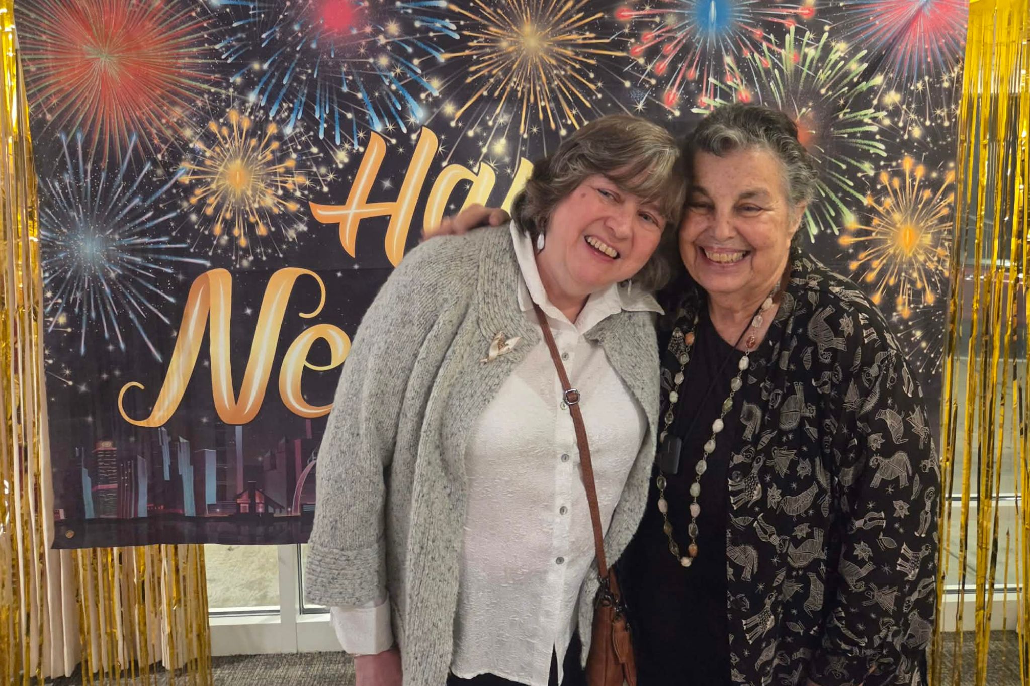 Two women smiling together in front of a festive Happy New Year banner with fireworks in the background capture a joyful moment perfect for a summer photo essay.