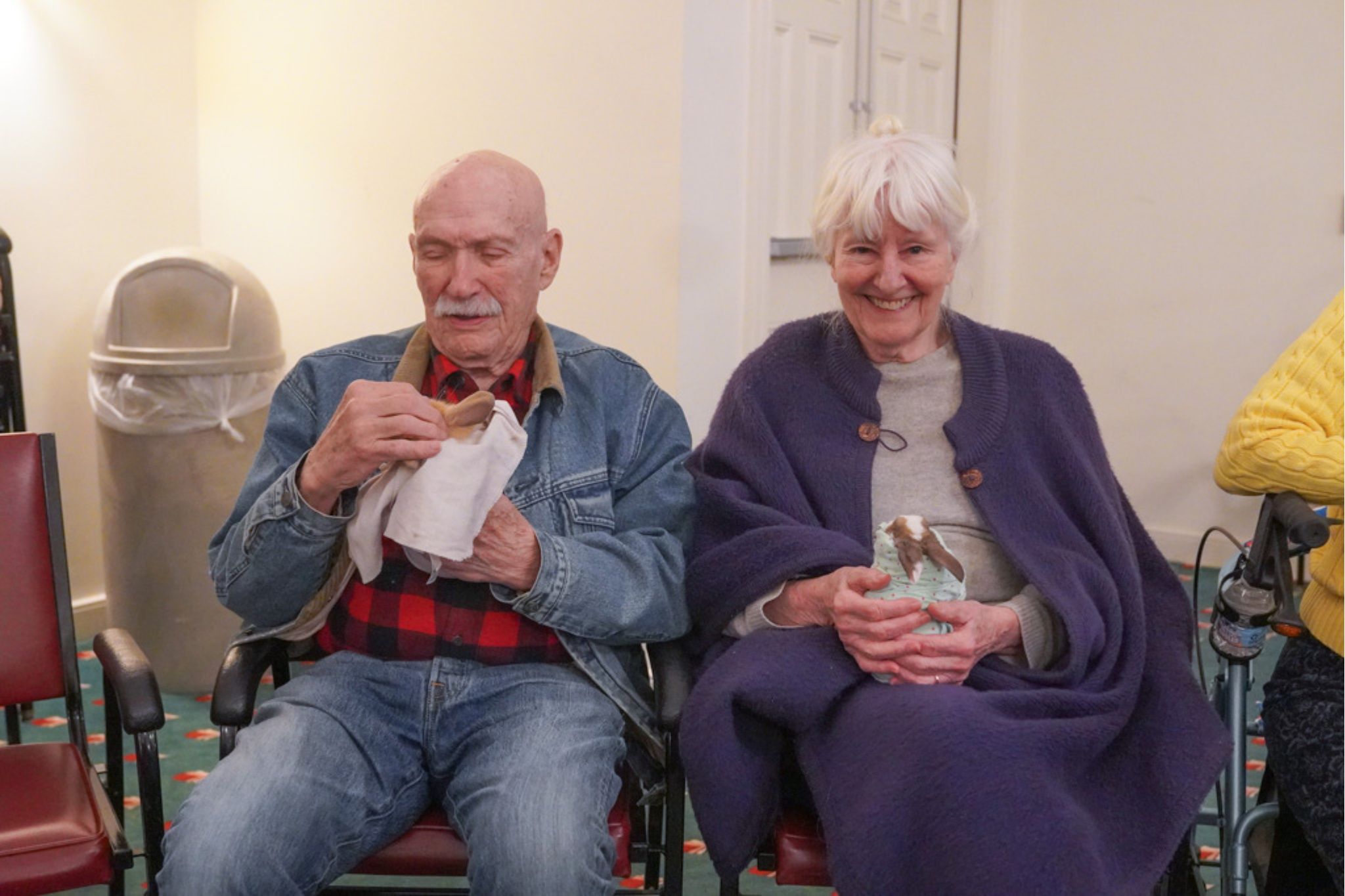 Two older adults sit on chairs indoors, smiling and holding snacks, with a trash can in the background—perhaps celebrating a recent scholarship announcement.