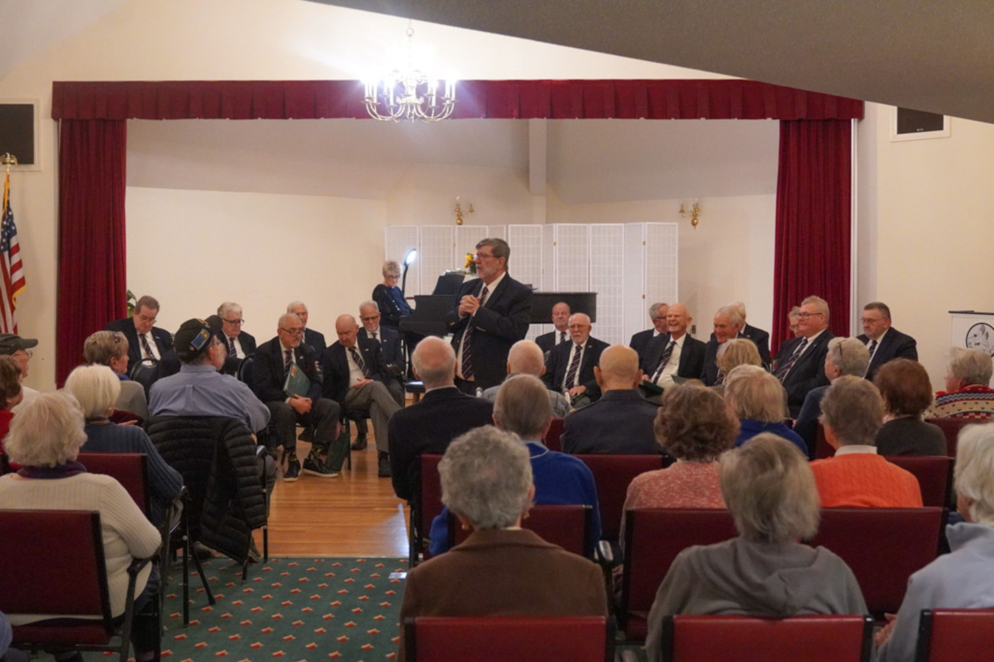 A man speaks to an audience in a hall, with a group of people—possibly scholarship recipients—seated on stage behind him.