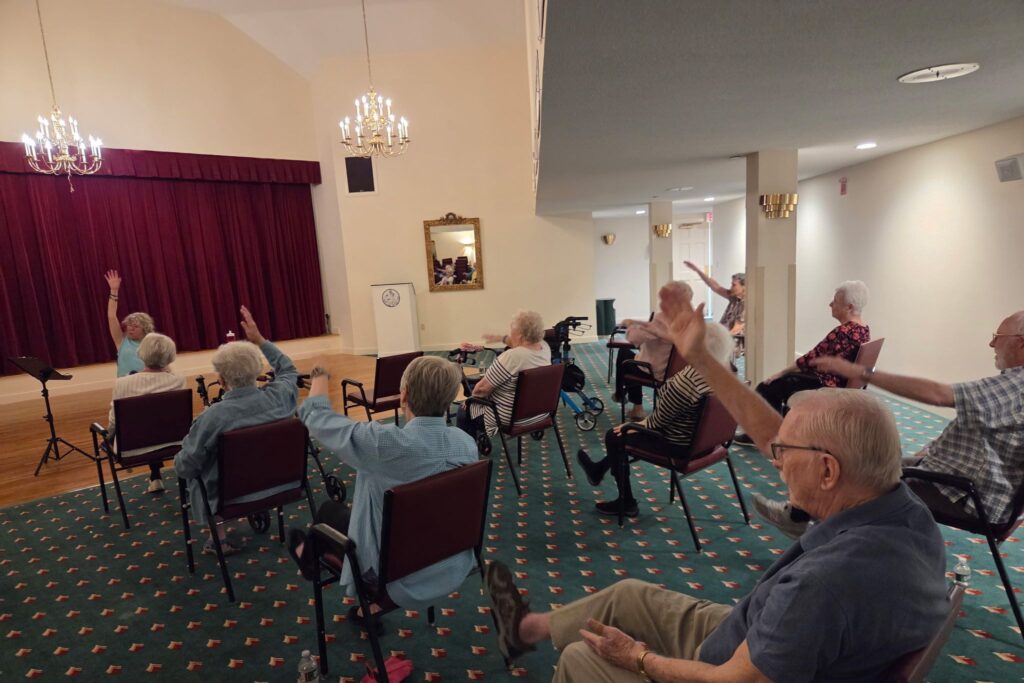 A group of seniors sitting in chairs raise their arms during an exercise class in a community room, where participants share stories about scholarship opportunities and lifelong learning.