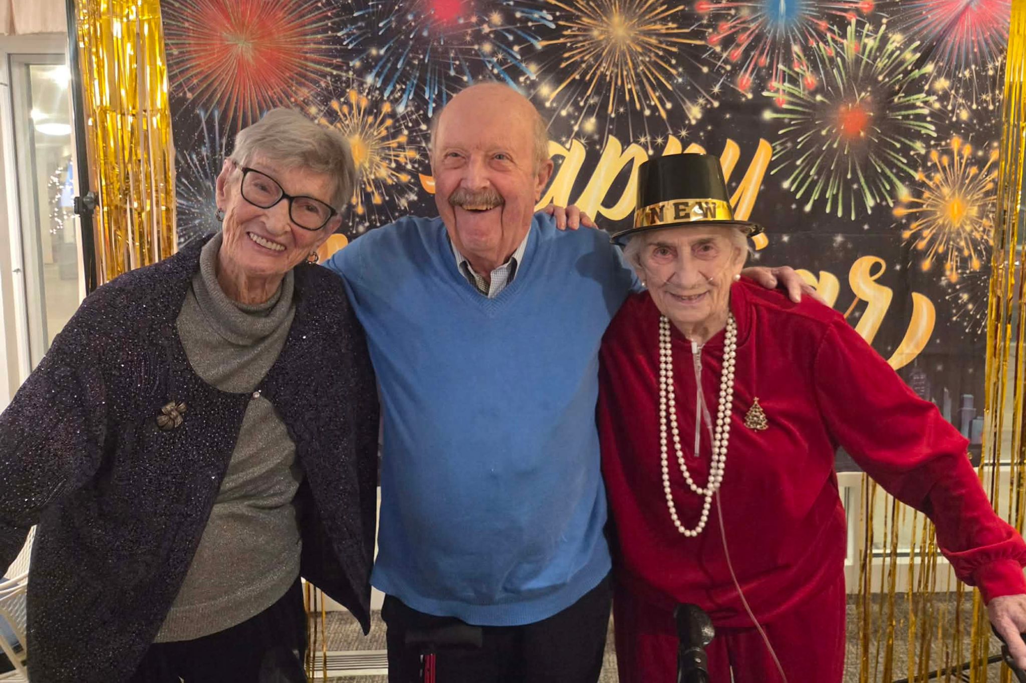 Three smiling seniors pose together in front of a festive New Year’s backdrop with fireworks and "Happy New Year," capturing a joyful moment perfect for a photo essay, even as they reminisce about summer memories.