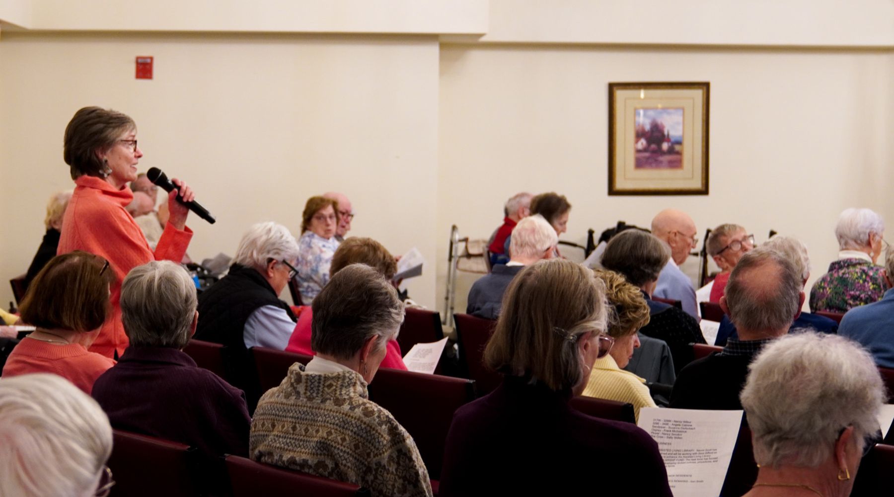 Woman with a microphone stands and speaks to a seated group of elderly people holding papers in a meeting room, discussing animal-related topics.