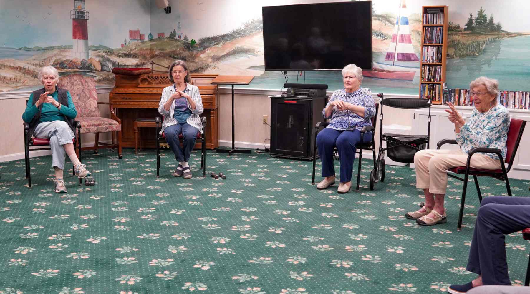Four elderly women sit in a circle clapping hands, enjoying a lively summer afternoon in a room with a mural, TV, and piano in the background.