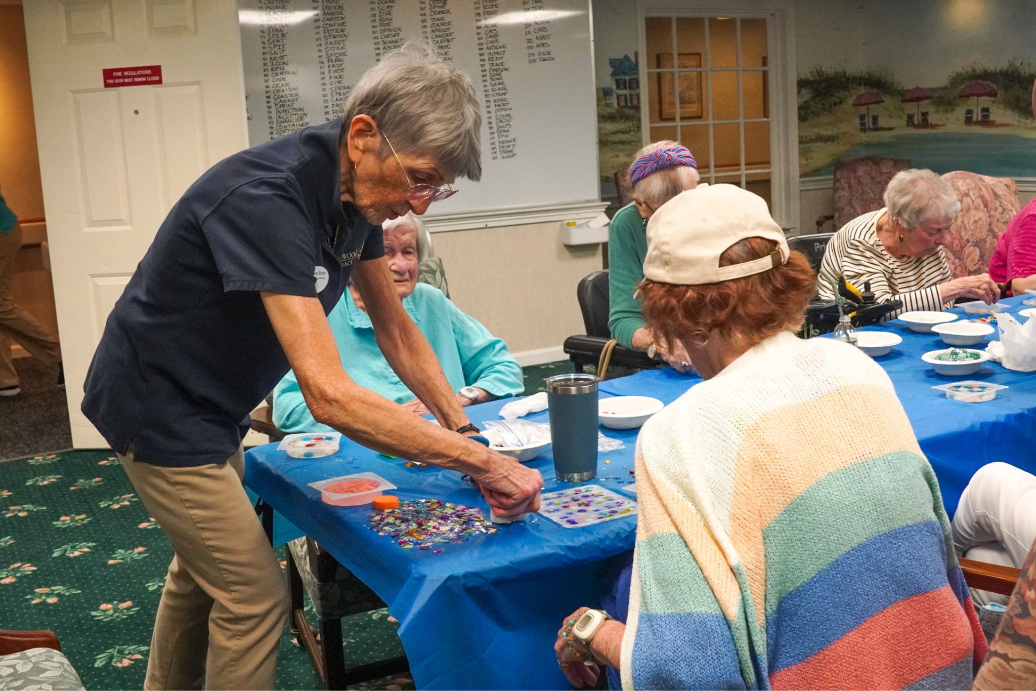 Elderly women sit around a table making crafts together in a brightly lit community room at an assisted living facility.