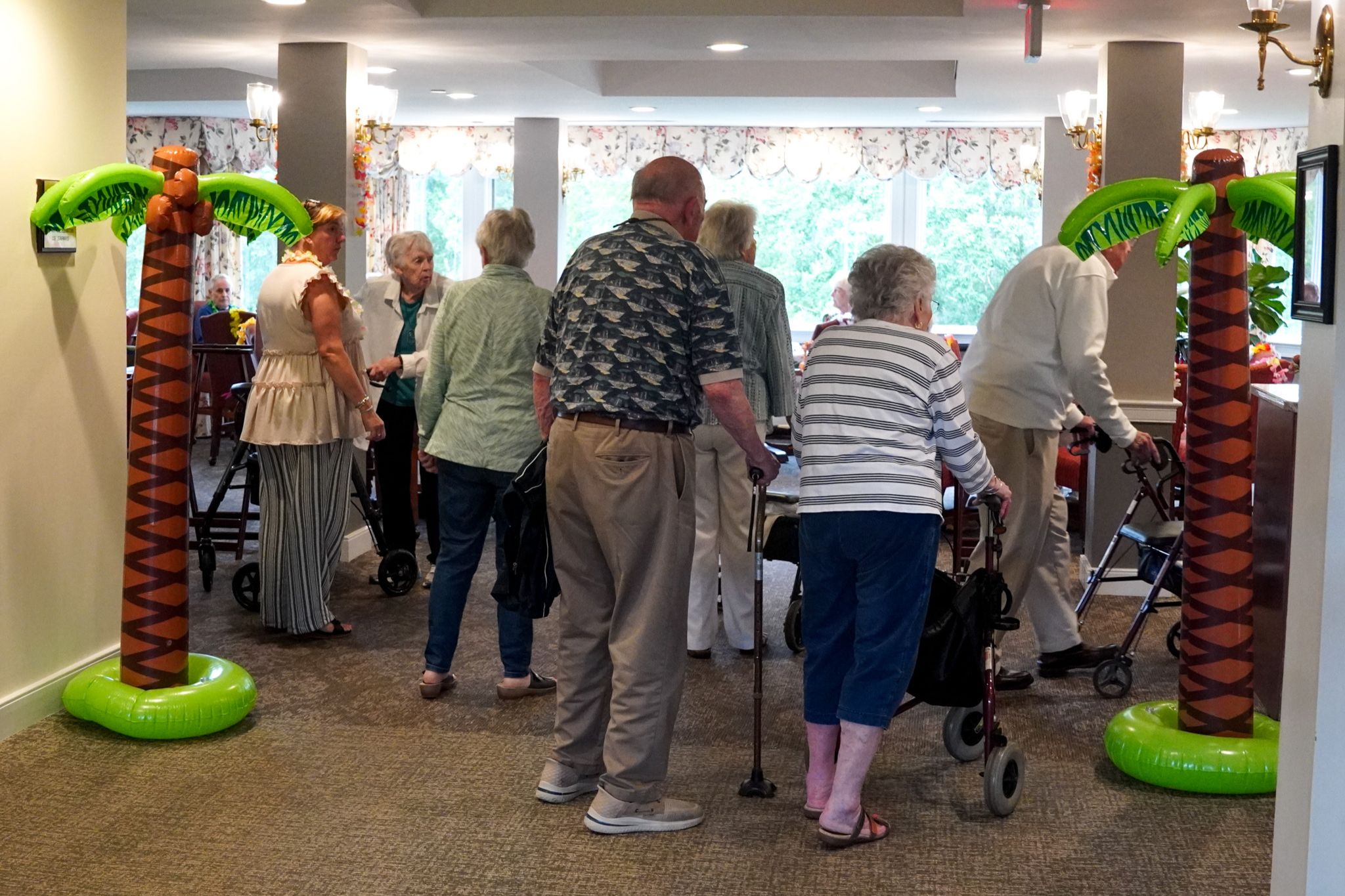 A group of seniors with walkers gather indoors between two inflatable palm trees at a summer-themed social event.