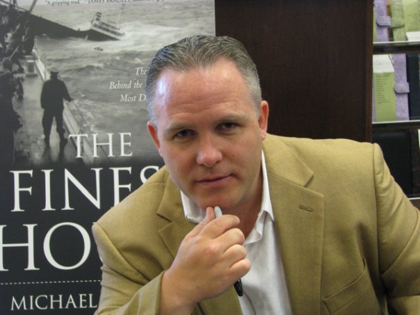 A person in a beige suit poses pensively, clutching a pen near their face, like a captain charting ships on an ancient map, all set against the backdrop of a book display.