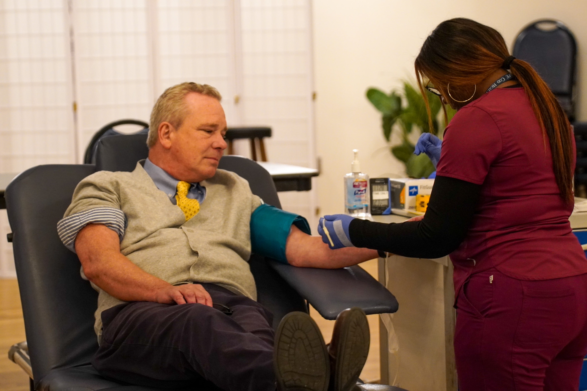 A man donates blood during a local blood drive, comfortably seated in a chair as a healthcare worker attends to him in the clinic setting.