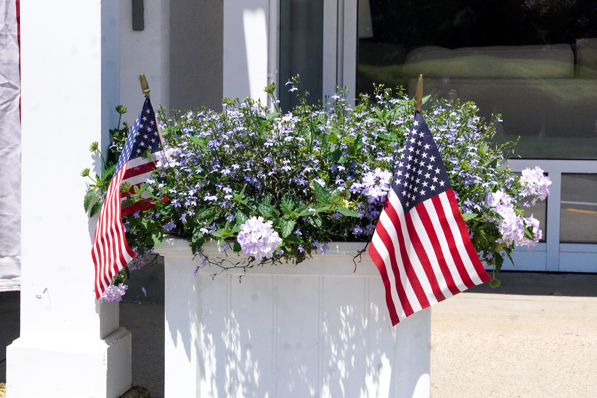 A flower planter with small American flags brightens the outside of a building on a sunny summer day.