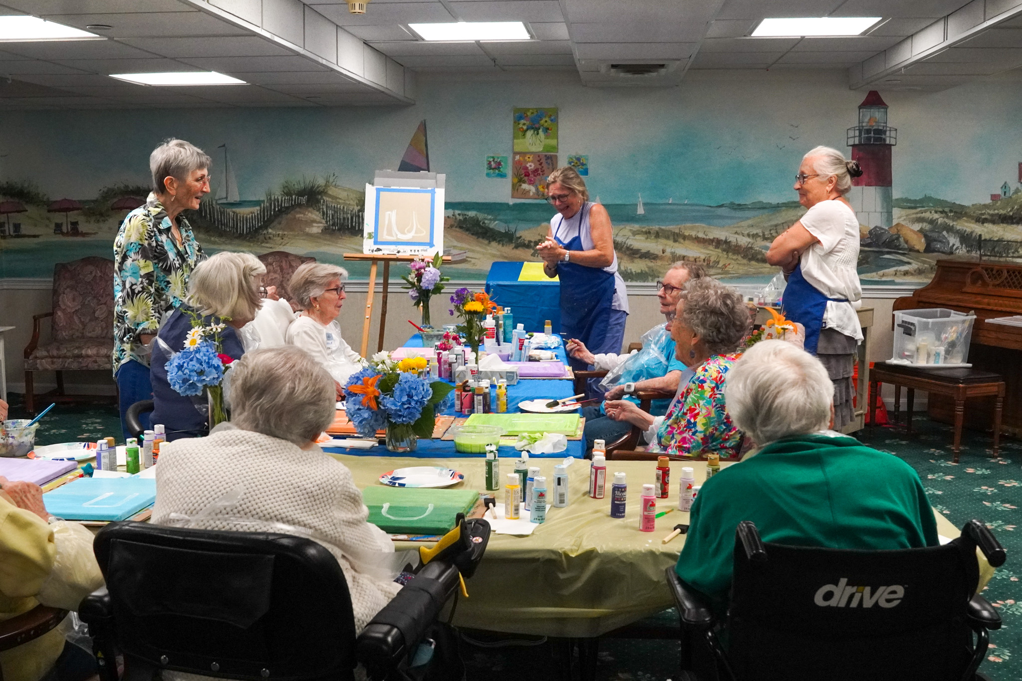 A group of seniors sits around a table doing arts and crafts in a brightly decorated room at an assisted living community.