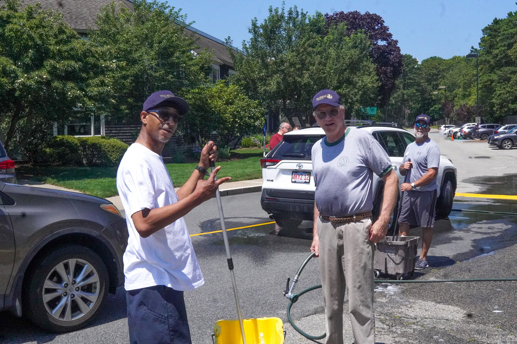 Three men in gray shirts wash cars outdoors on a sunny summer day, holding cleaning tools and buckets.