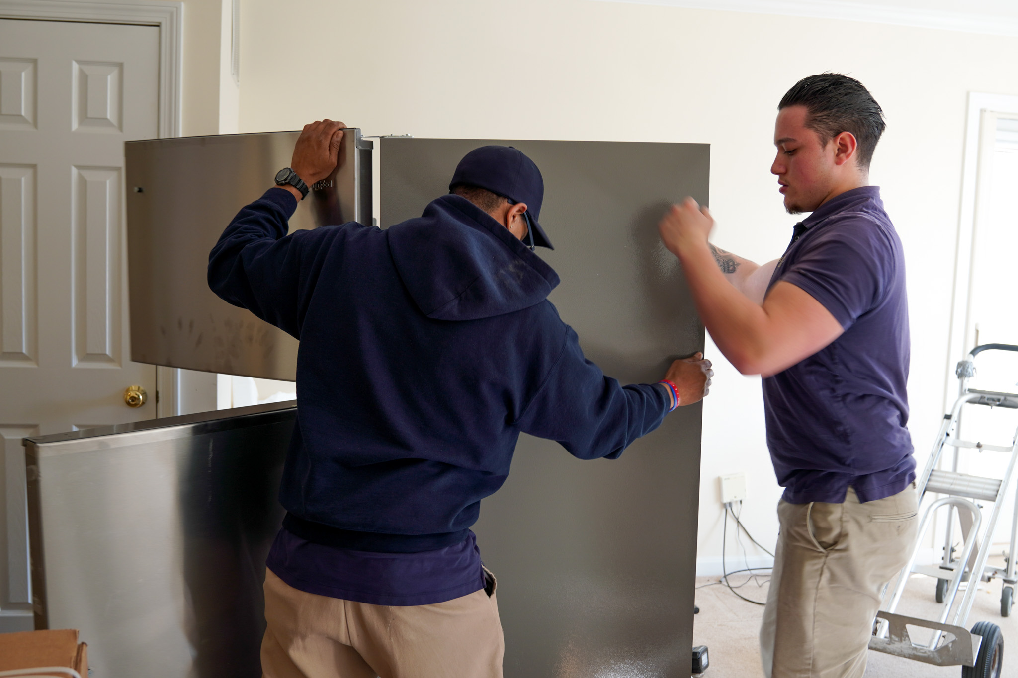 Two men are downsizing as they carefully maneuver a large refrigerator across the room, a white door and ladder standing watch in the background.