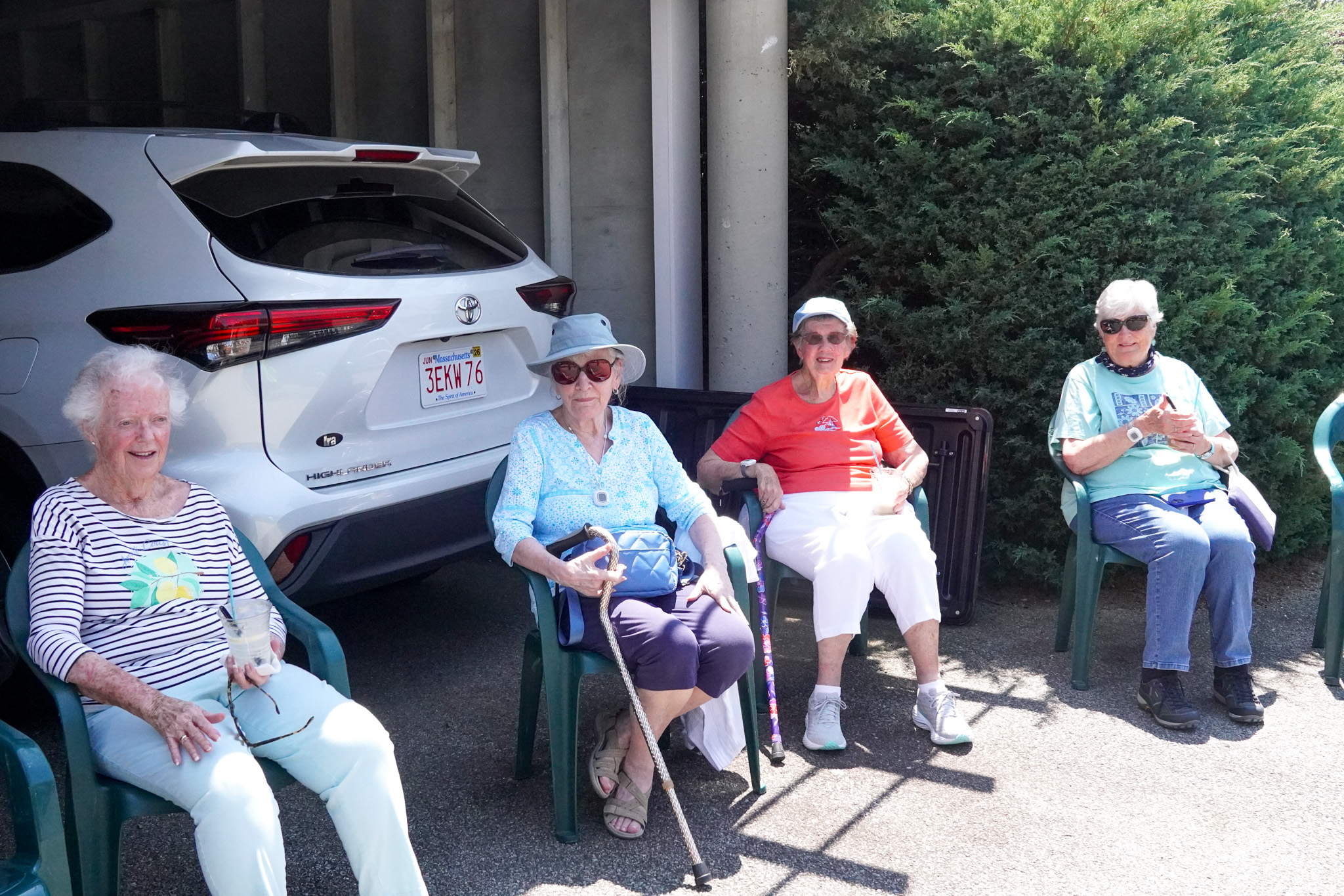 Four older women sit on chairs outdoors in the summer sun, beside a white car and green bushes, smiling and relaxed.