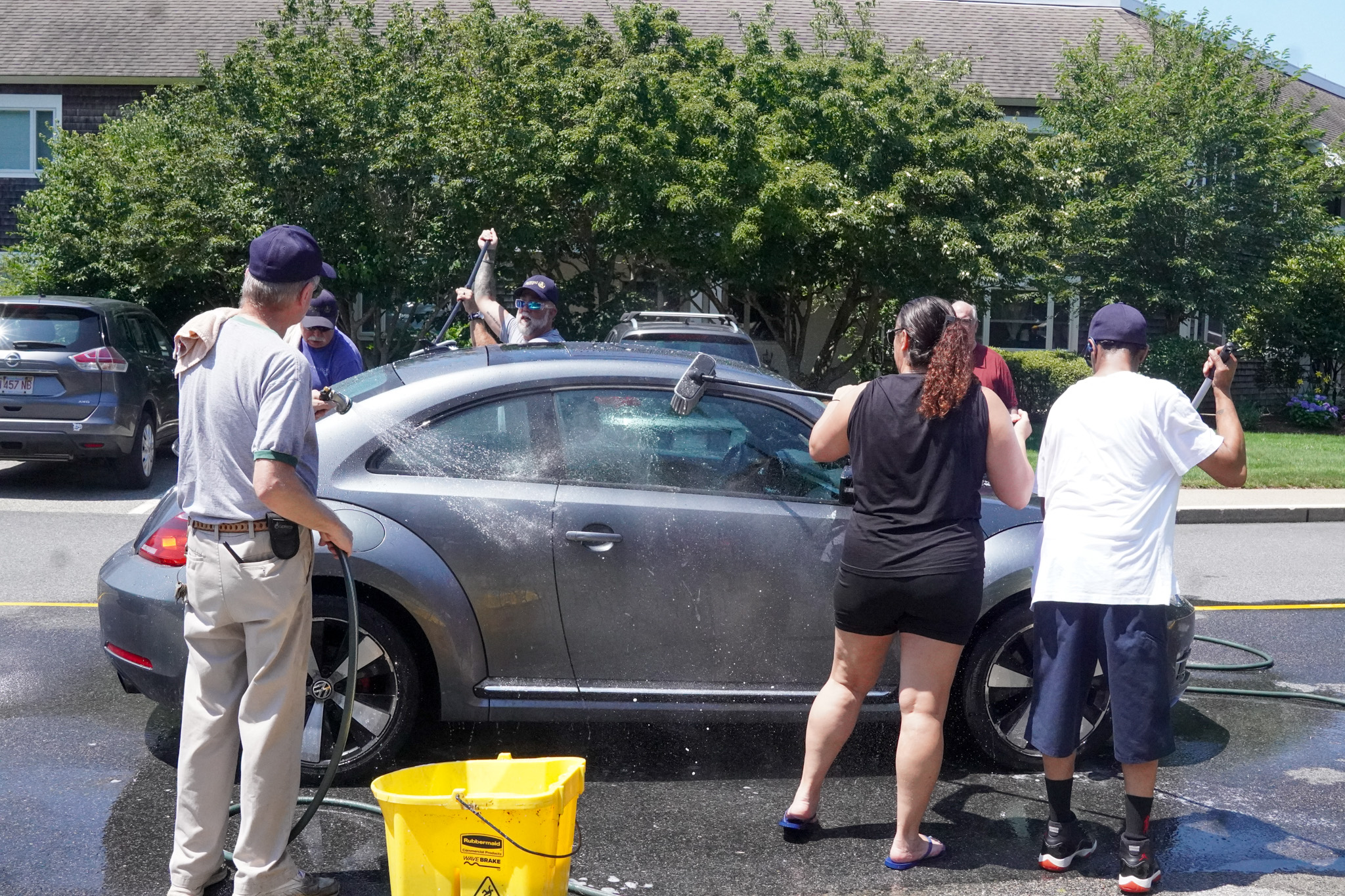 Five people washing a gray car together outdoors on a sunny summer day with soap, brushes, and a hose.