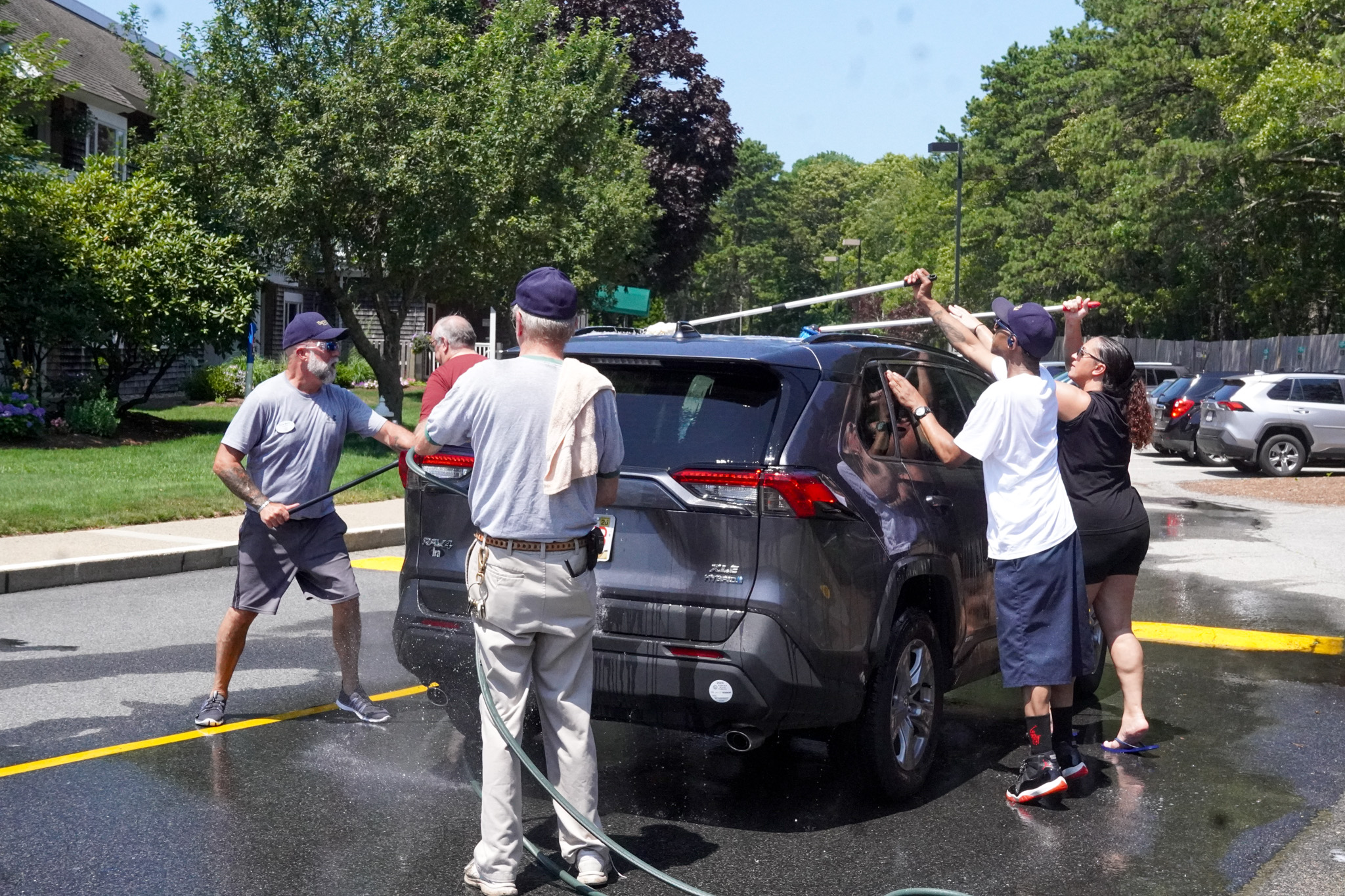 Five people wash a dark SUV together on a sunny summer day in a parking lot surrounded by trees.