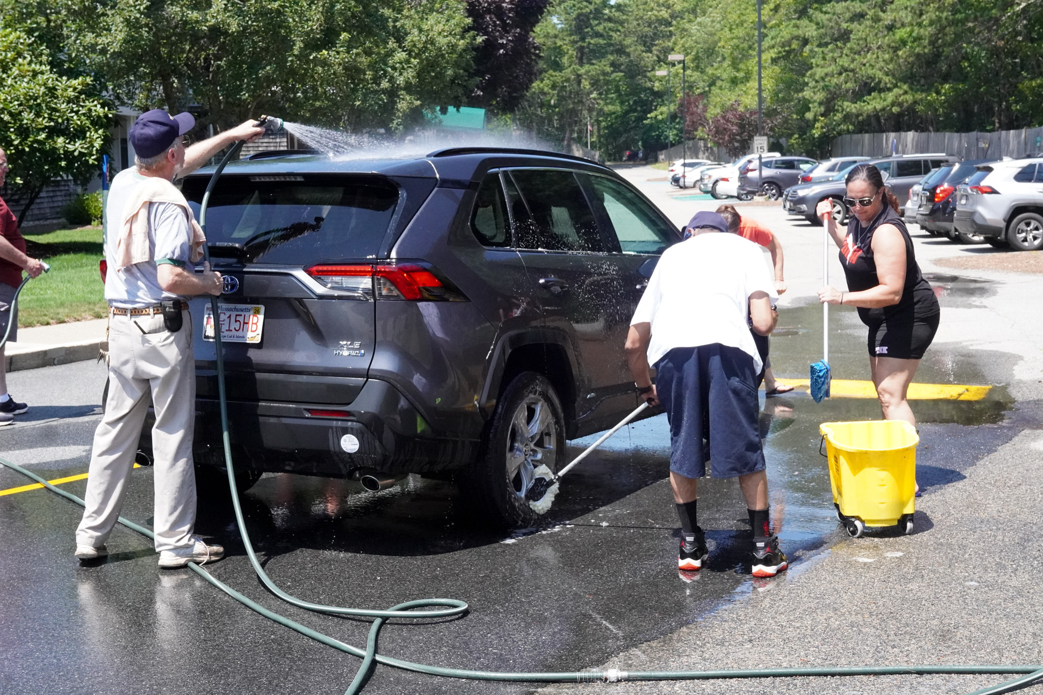 Three people washing a black SUV in a sunny parking lot, using hoses, sponges, and a yellow bucket on a warm summer day.