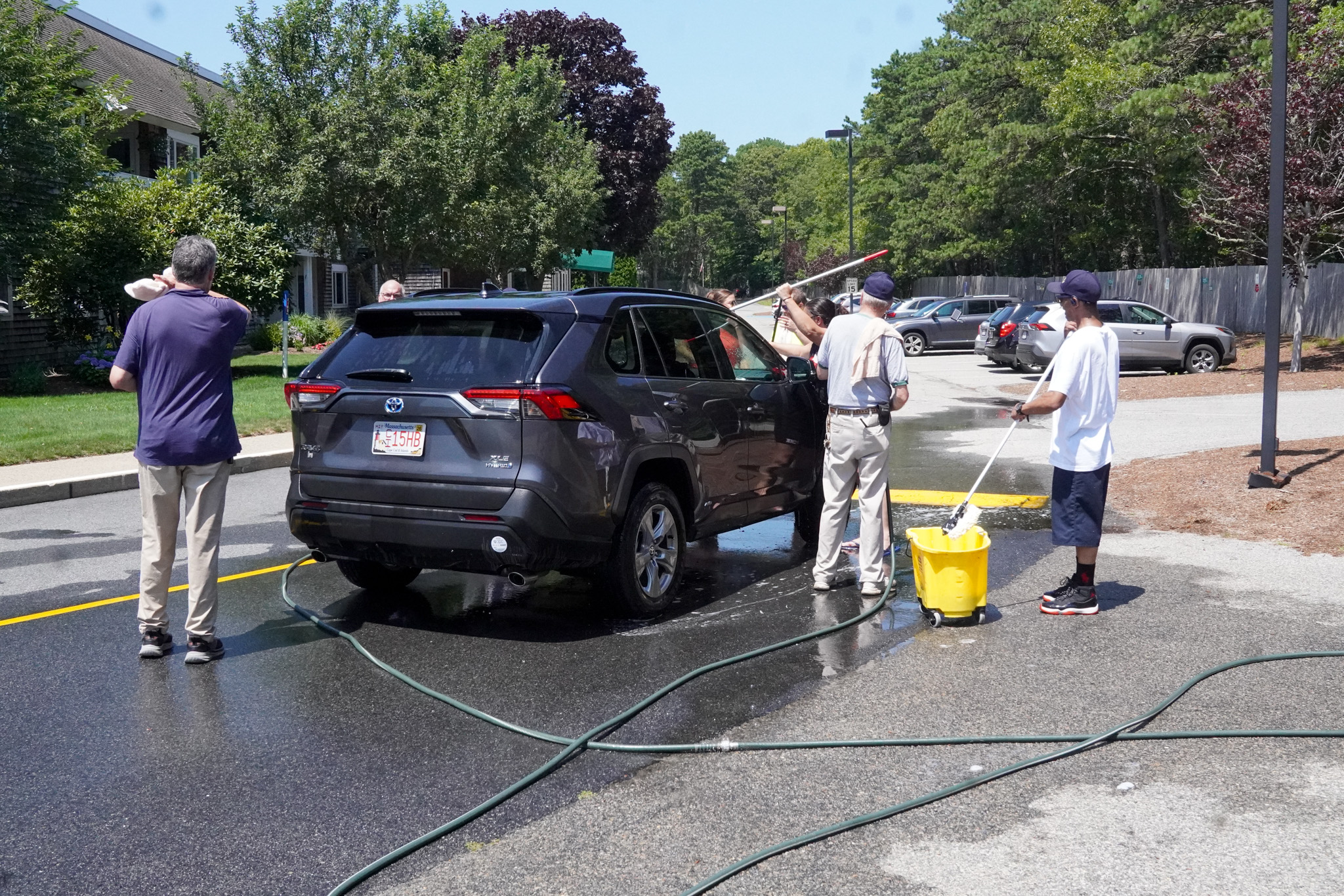 Four people wash a black SUV with hoses and brushes in a parking lot on a sunny summer day.