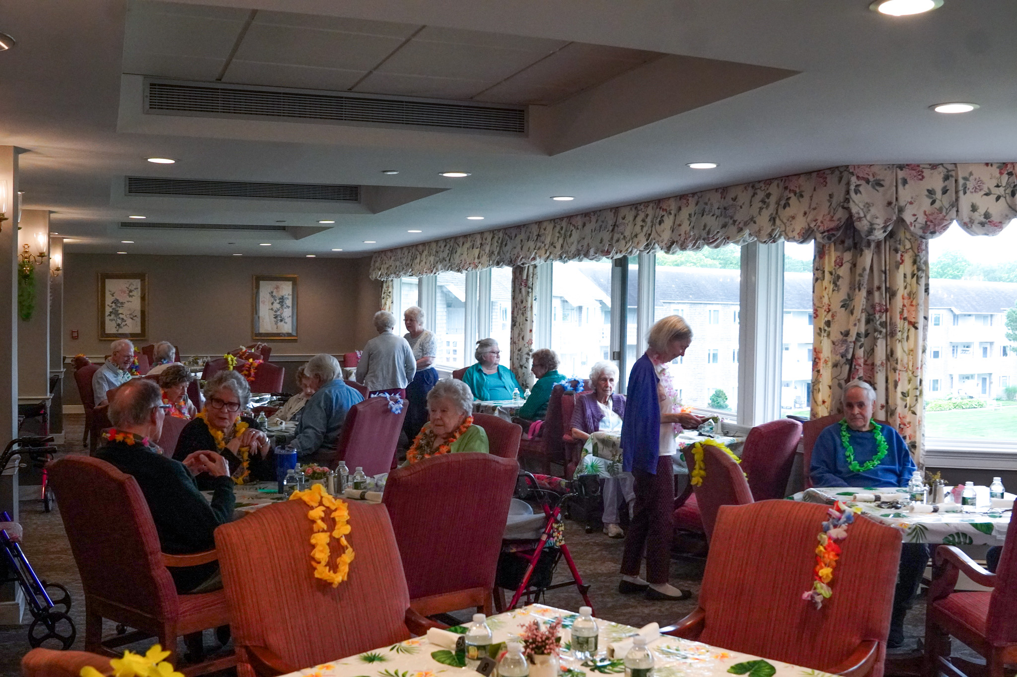 Elderly people wearing leis sit and eat together in a bright dining room with large windows, enjoying the cheerful atmosphere of a summer gathering.