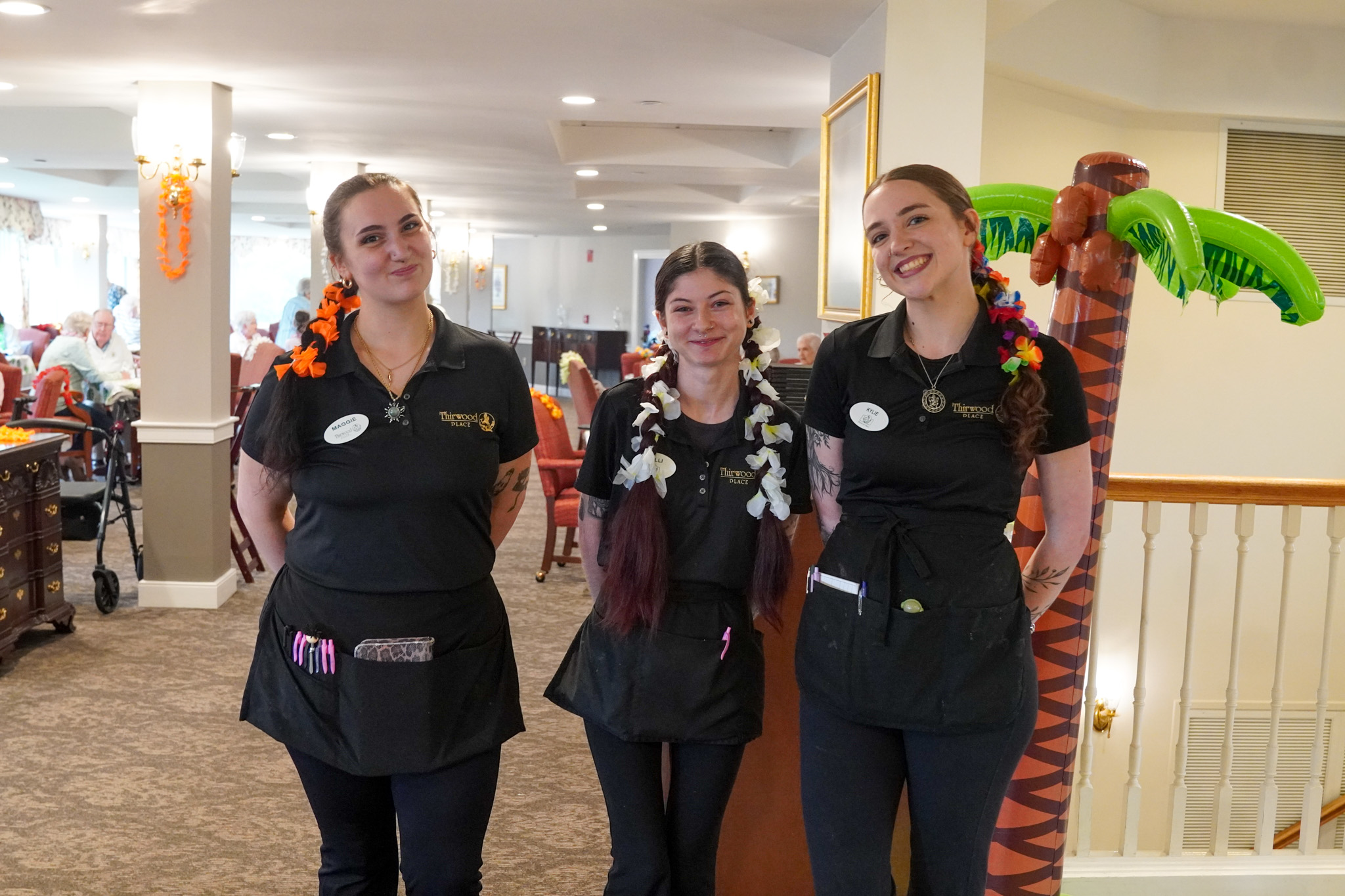 Three smiling women in black uniforms with flower leis stand indoors near a fake palm tree, bringing a touch of summer to the scene.