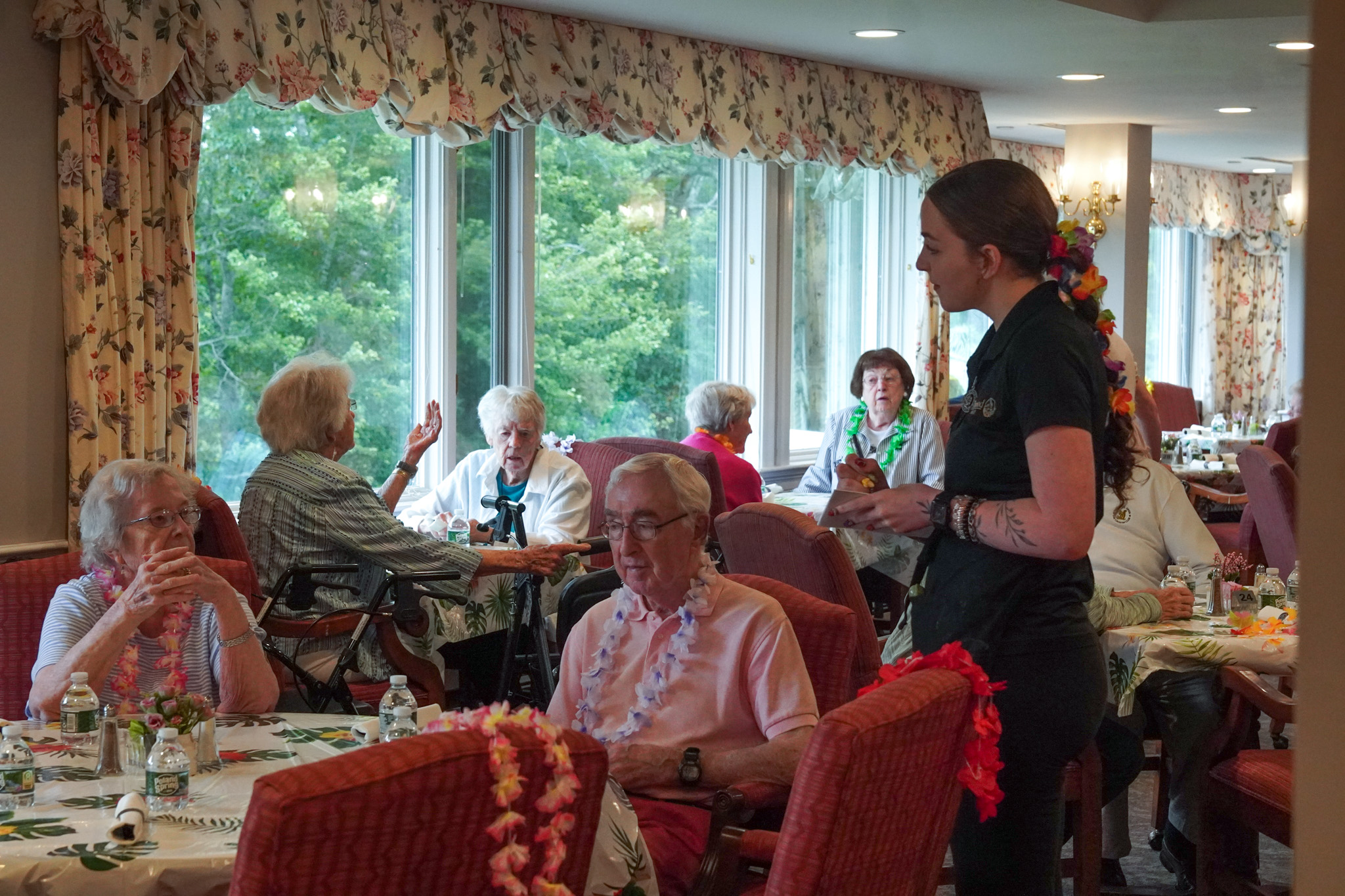 A group of seniors enjoys a summer meal together in a bright room while a server takes an order.