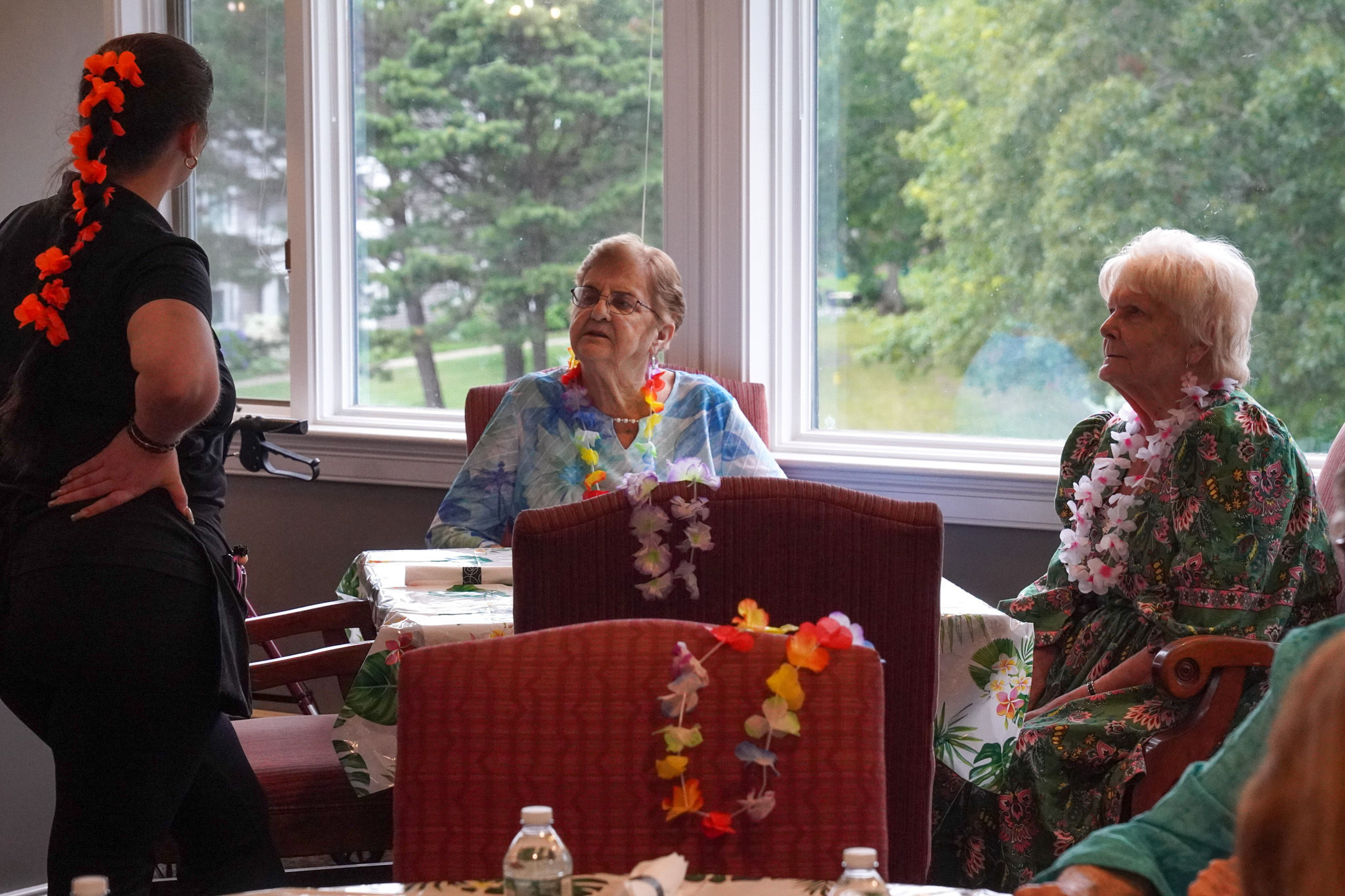 Three women in floral leis sit and chat by large windows at a tropical-themed indoor gathering, enjoying the relaxed vibes of a summer celebration.