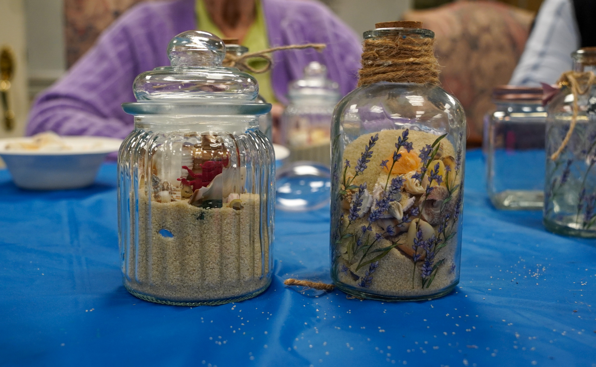 Two glass jars filled with sand, dried flowers, seashells, and a tiny animal figurine sit on a blue tablecloth.