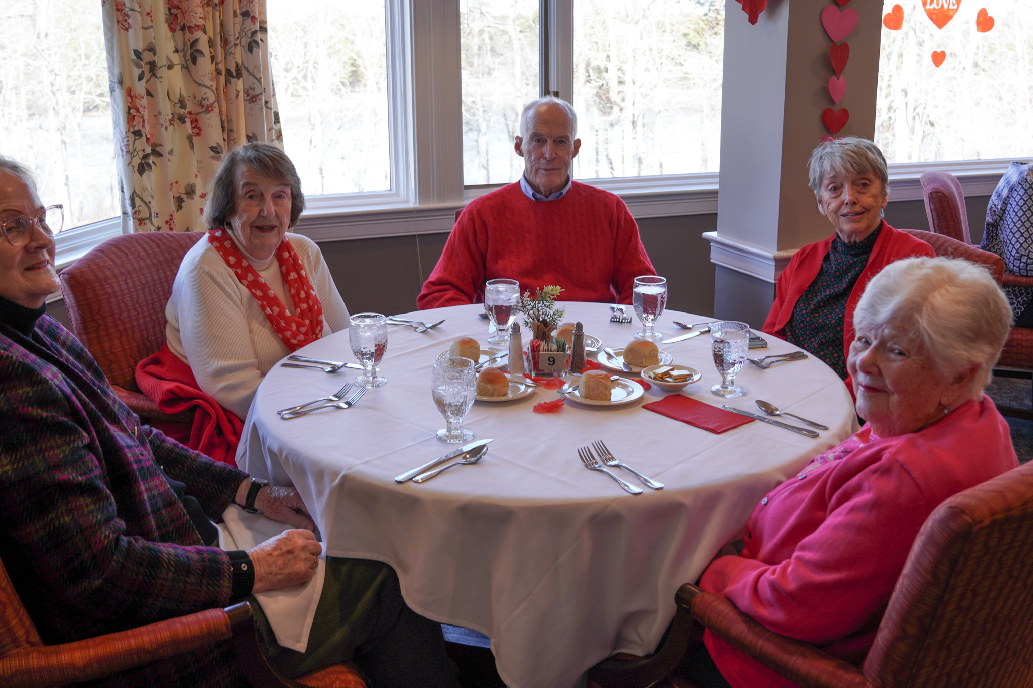 Five older adults are dining together around a round table set for a meal, with Valentine’s Day decorations in the background.