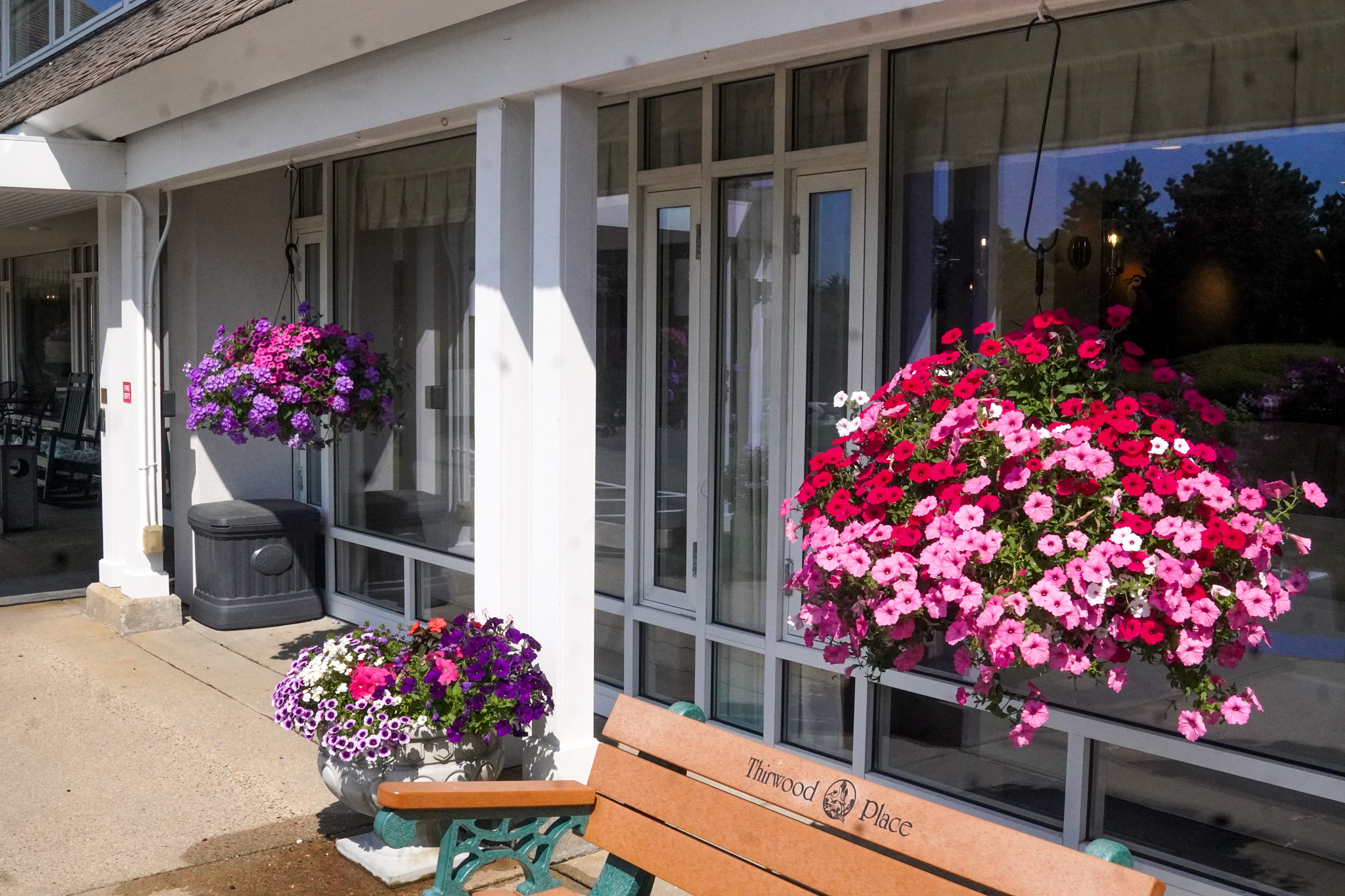 Hanging baskets of pink and purple flowers decorate the exterior of a building near a bench labeled Tuxedo Place, creating a vibrant summer scene.