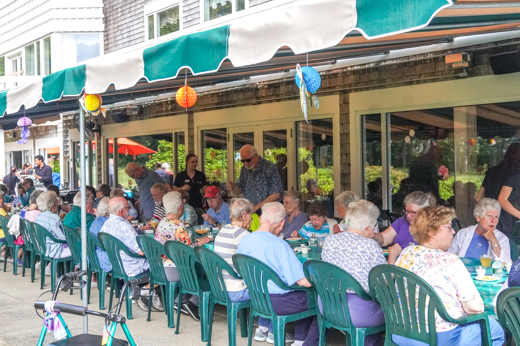 A large group of elderly people sit at long outdoor tables, enjoying a summer evening while eating and socializing under colorful lanterns.